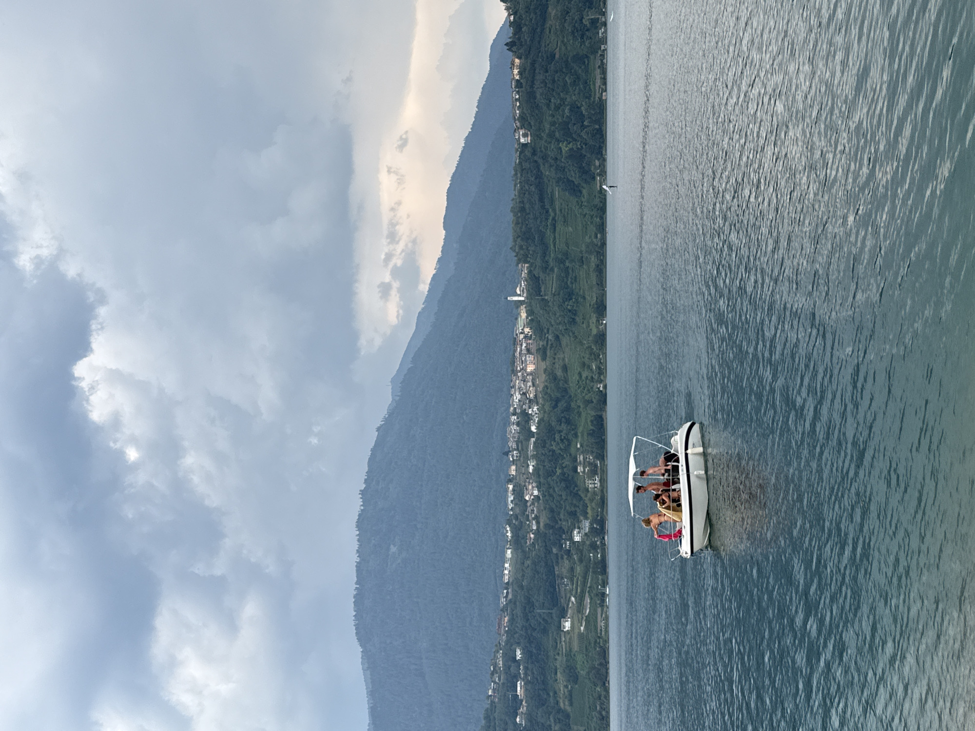In the city of Calceranica al Lago, Italy, a white motorboat with a canopy is navigating across a wide, dark blue lake. Onboard are four individuals, two women and two men. One woman is standing and holding a pink towel. A second woman is seated. The two men are also seated. In the background, a densely populated shoreline is visible, with buildings scattered across a green, tree-covered hillside. A tall, slender white structure, possibly a church steeple, stands out amongst the buildings. Beyond the shoreline, imposing, dark green mountains rise up, their peaks partially obscured by heavy, grey, cloudy skies. The water exhibits a slight ripple pattern, and a faint reflection of the boat is visible on its surface.