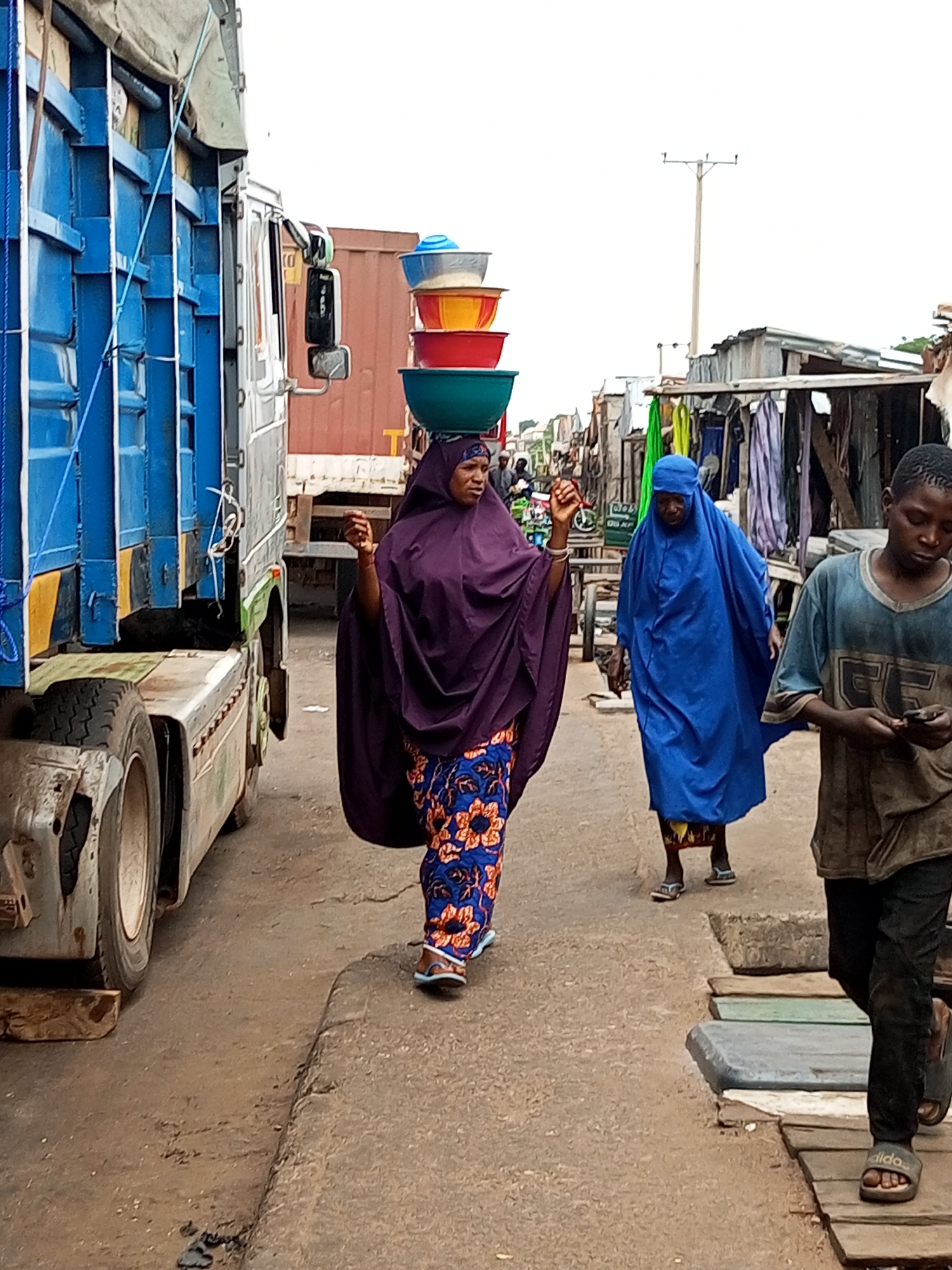 The image depicts a street scene in the city of Gombe, Nigeria, likely within a market area, given the presence of stalls and a truck laden with goods. In the foreground, a person wearing a purple hijab and a patterned dress carries a stack of colorful plastic bowls on their head. To their right, another person in a bright blue hijab and dress walks, and further to the right, a young person in a dark t-shirt and pants, carrying a phone, walks away from the camera. A large blue and silver truck occupies the left side of the frame, its side panels and a portion of its tires visible. The ground appears to be unpaved, with some wooden planks laid out in sections, suggesting a makeshift walkway or market stalls. In the background, a utility pole stands tall against a bright, overcast sky. The overall impression is one of bustling daily activity in a vibrant urban environment.