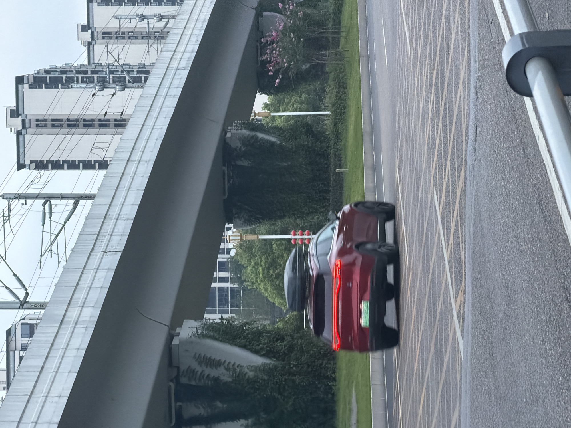 A red SUV with a roof cargo box is driving on a road in Jiangjin, China. The car is blurry due to motion. The road has yellow and white grid markings. In the background, there is an elevated structure, possibly a bridge or railway overpass, with overhead wires. Behind the elevated structure, there are several tall, modern buildings and lush green trees. The sky is overcast.