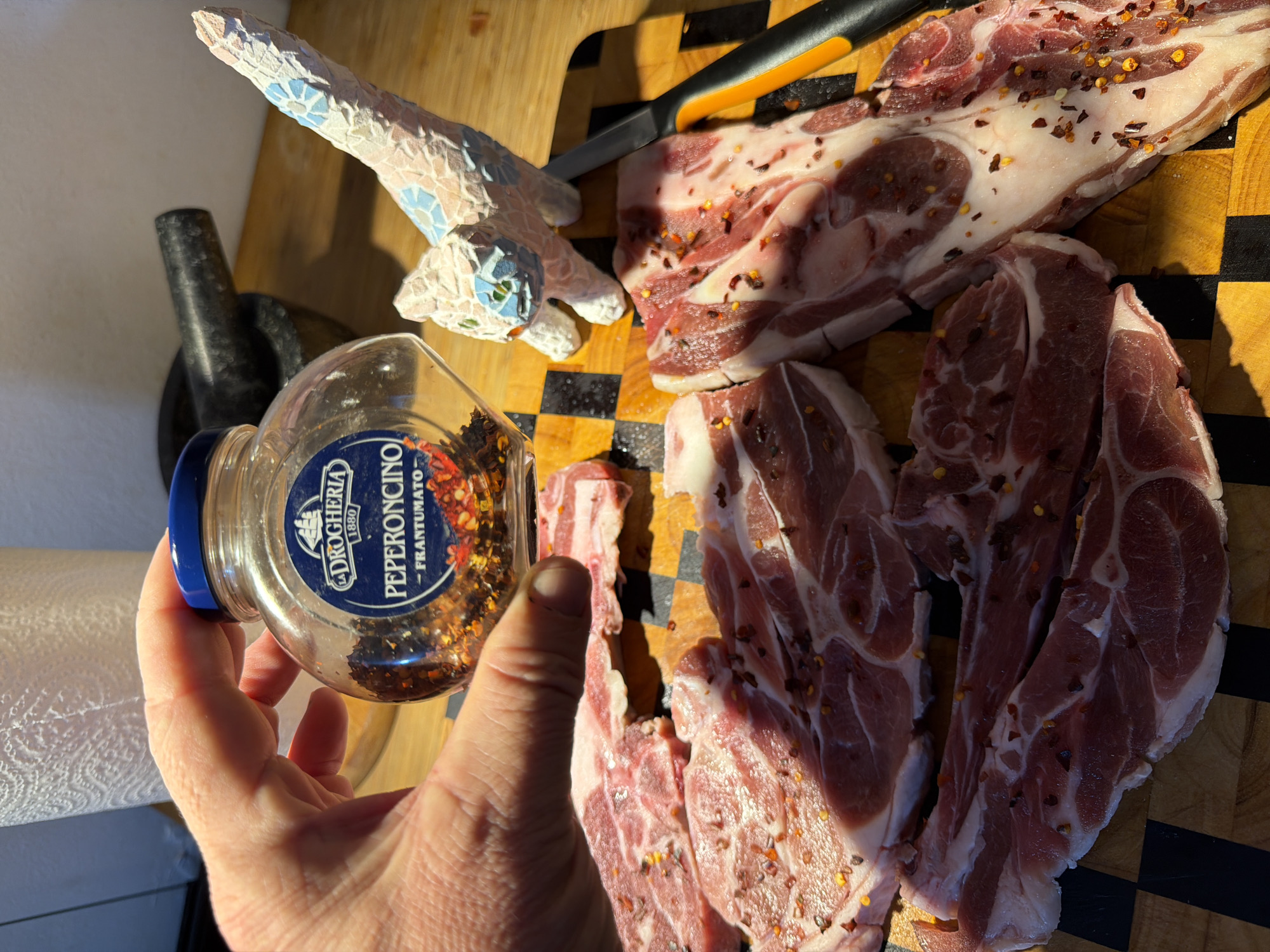 A close-up, overhead shot shows raw meat seasoned with red pepper flakes on a cutting board. In the foreground, a hand holds a jar of "Peperoncino Frantumato" from "La Droghesia 1880". To the right of the meat, a mosaic cat figurine stands next to a knife. In the background, a mortar and pestle sits on a wooden surface, with a roll of paper towels to the left. The scene appears to be a kitchen preparation area. This image was taken in Even Yehuda, Israel.