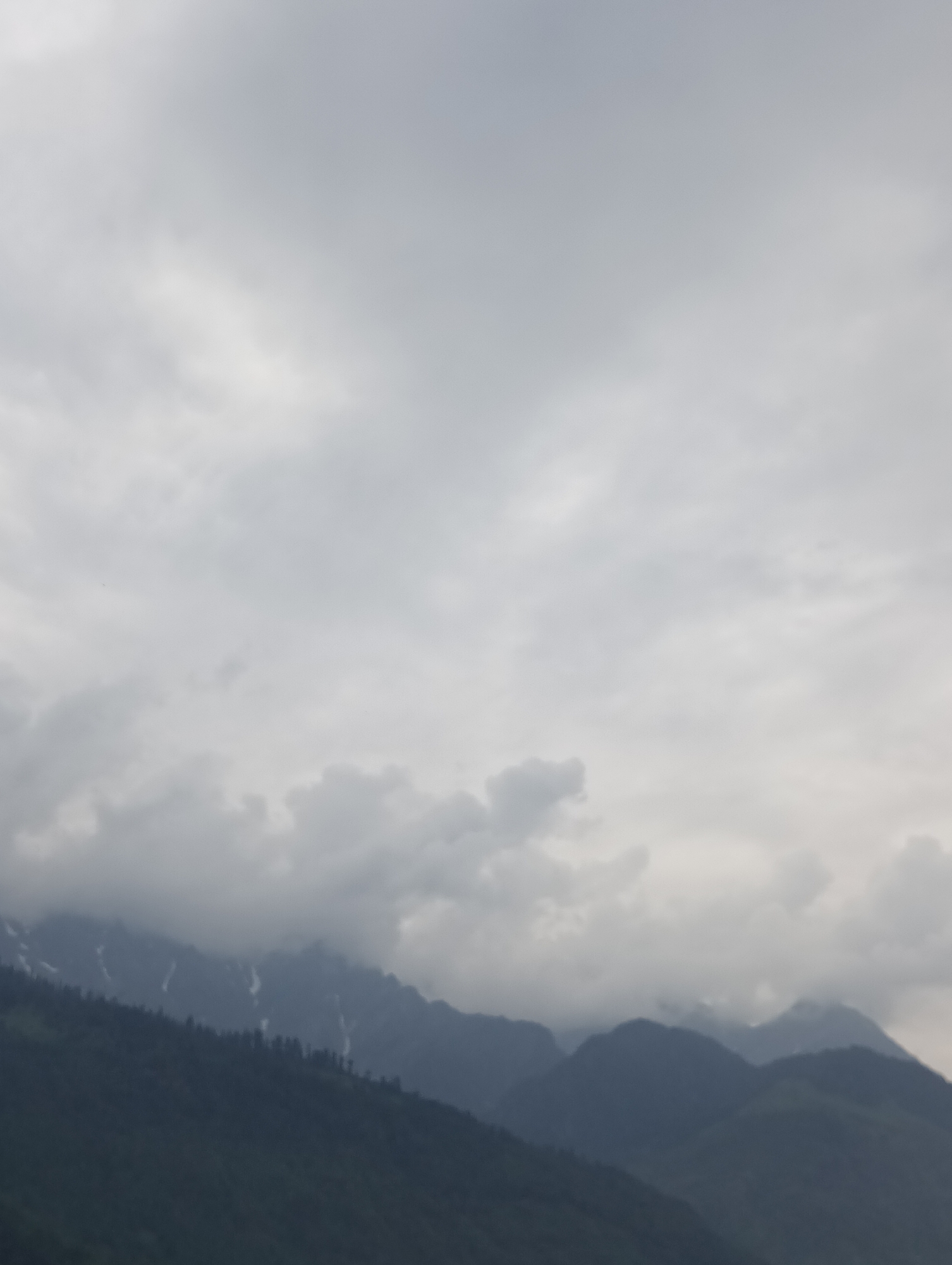 The image depicts a hazy, overcast sky above a range of dark, silhouetted mountains. The clouds are thick and grey, obscuring the sky and suggesting a dull or rainy day. The mountain slopes are covered in deep green vegetation, with some patches of white snow or frost visible higher up. The overall atmosphere is muted and somewhat somber due to the lack of direct sunlight and the presence of fog or mist. The scene appears to be a natural landscape, likely in a mountainous region, given the prominence of the peaks. There are no people or man-made objects visible, focusing the attention entirely on the natural environment. The time of day is indistinct due to the cloud cover, but it appears to be daytime. The location cues, combined with the visual elements, suggest a scene from Vashisht, India, a region known for its mountainous terrain and often cloudy weather. The mood evoked is one of natural grandeur, perhaps tinged with a sense of mystery or tranquility brought on by the atmospheric conditions.