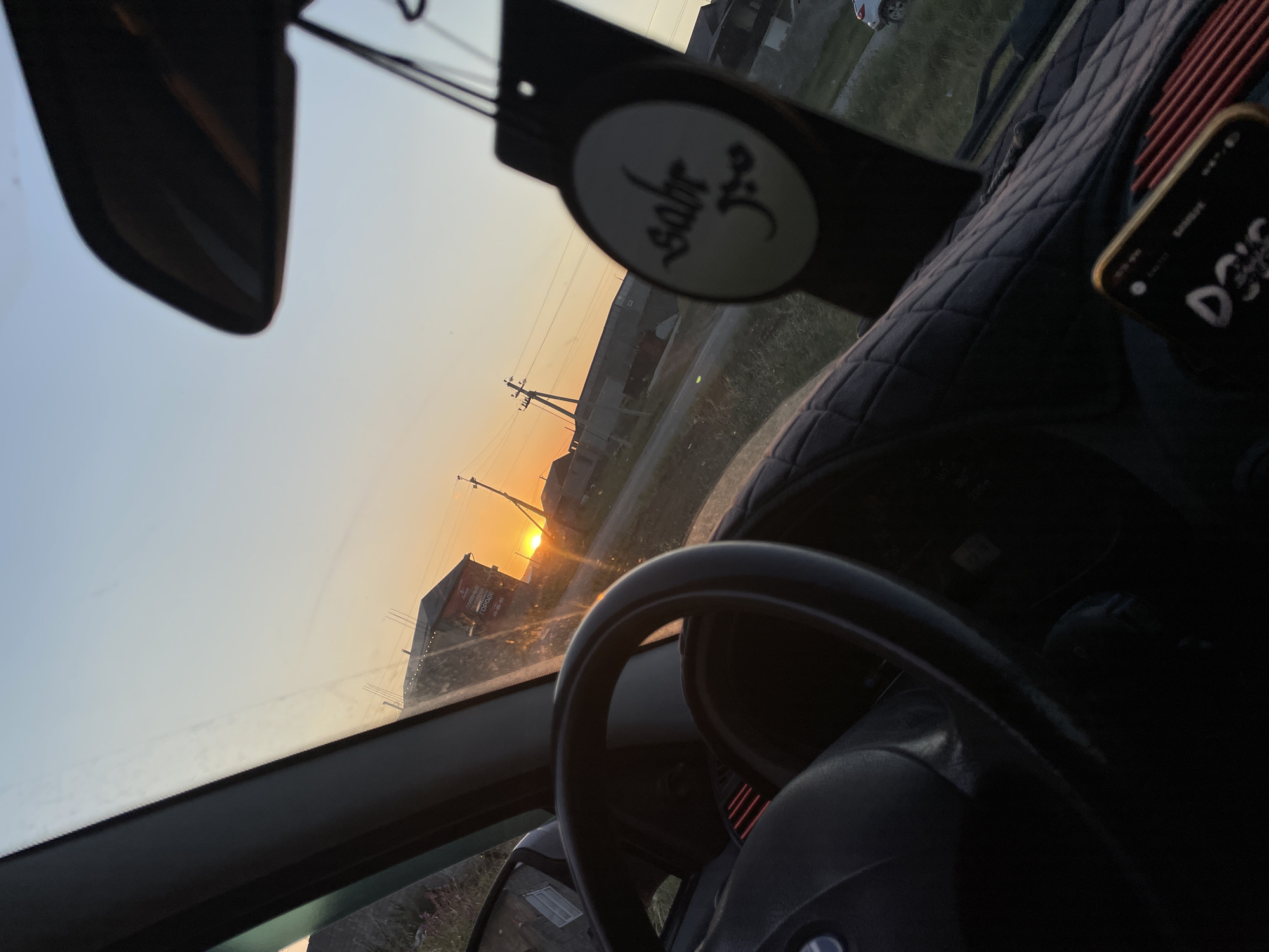 The image provides an interior view from a car, looking out the windshield at a sunset. The scene is set in a rural or semi-rural area, with buildings and power lines visible against a soft orange and blue sky. The sun is low on the horizon, casting a warm glow.
Dominant objects within the car include the steering wheel, a rearview mirror, and a dashboard. A patterned car seat cover is visible on the passenger side. Hanging from the rearview mirror is an air freshener with an oval tag that has some script written on it, appearing to be in Arabic or a similar script. A smartphone is also visible on the dashboard, showing a lock screen with some text.
The time of day is clearly sunset, given the low angle of the sun and the warm, diffused light. The weather appears to be clear. The location cues, such as the buildings and power lines, suggest an outdoor setting in a developed, though possibly sparse, area. There are no people visible in the image. The overall mood is serene and reflective, perhaps conveying a sense of journey or transition associated with the end of the day.