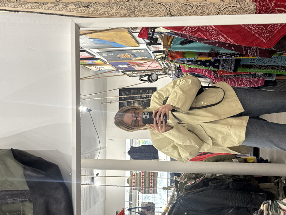 The image captures a mirror selfie taken by a female subject inside a retail establishment located in the city of Unknown, Unknown. The individual, possessing blonde hair and wearing dark-framed glasses, holds a dark-colored smartphone in front of her face, partially obscuring it. She is dressed in an oversized light-colored jacket and blue jeans, with a black strap, possibly from a shoulder bag, visible over her right shoulder. The mirror's reflection reveals a retail space characterized by numerous clothing racks filled with garments on both sides of the subject. To her right, a prominent vertical display of colorful patterned bandanas in red, blue, green, and black is visible. The wall behind the subject is covered with an assortment of framed artworks and posters, alongside a partial French sign. The ceiling shows exposed light fixtures, braided ropes, and a large green and black bag hanging. A pair of dark over-ear headphones hangs from a hook on the wall to the subject's right.