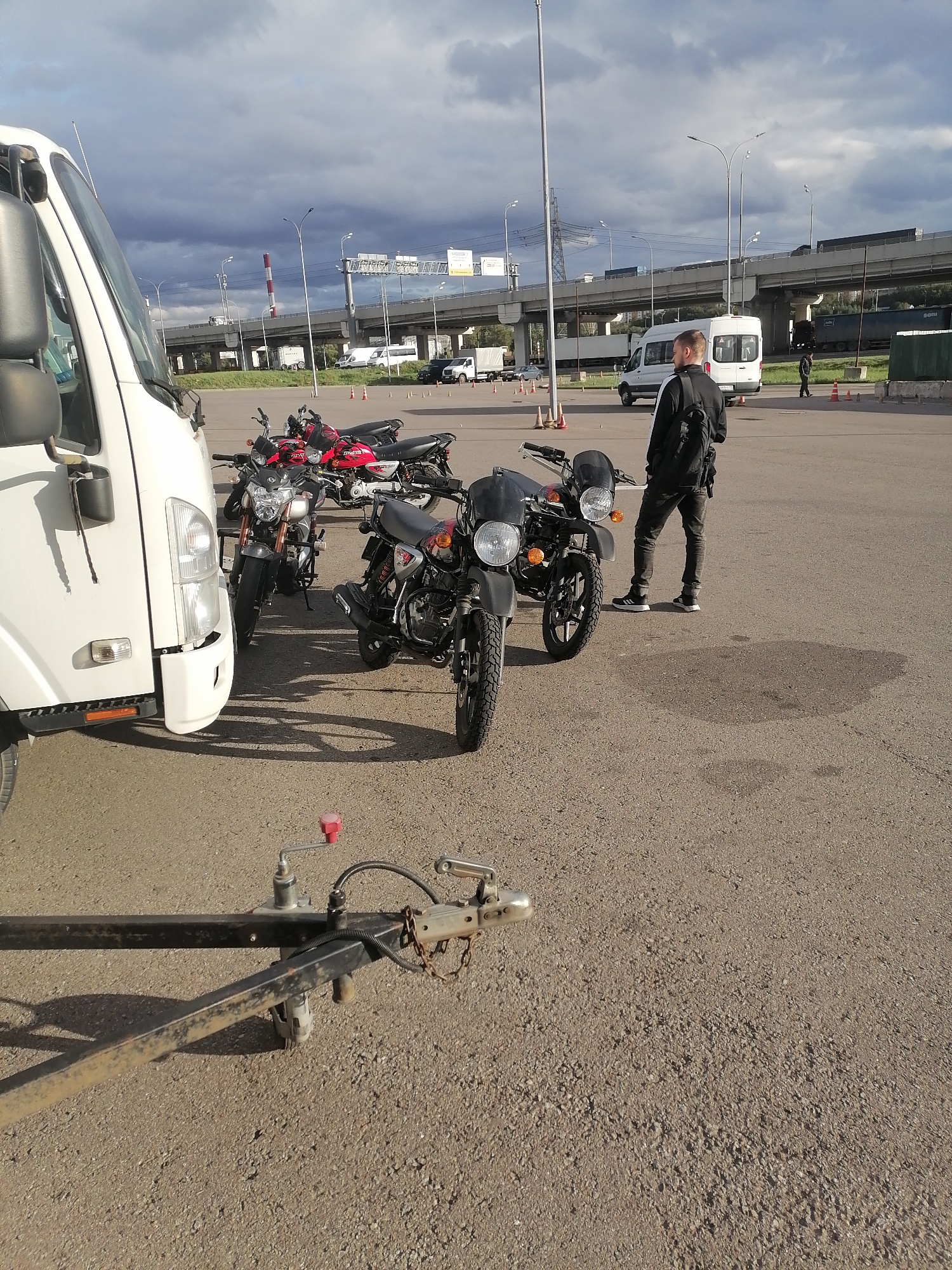 A man wearing a black jacket, black pants, and white sneakers stands in a parking lot, facing away from the camera. He has a black backpack on. To his left are two motorcycles, one red and one black. In front of the motorcycles is a white truck. To the right of the man is a trailer hitch. In the background, there are several vans and trucks, as well as an elevated highway with traffic. The sky is cloudy. The scene appears to be in Moscow, Russia.