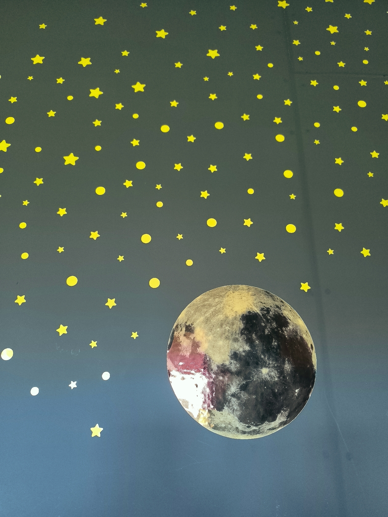 The image depicts a wall decorated with yellow stars and a large, realistic circular decal of the moon. The backdrop is a dark blue or teal color. The moon decal is prominently placed in the lower right portion of the frame, showing detailed craters and surface variations with a glossy finish. Scattered across the dark background are numerous small, bright yellow star-shaped decals and some yellow circular decals, resembling dots or smaller moons. A few white circular decals are also visible, mostly in the lower left section. The overall scene suggests a celestial theme, possibly in a child's room or a decorative space. There is no discernible location in the city of Almere, Netherlands, identifiable from the provided image.
