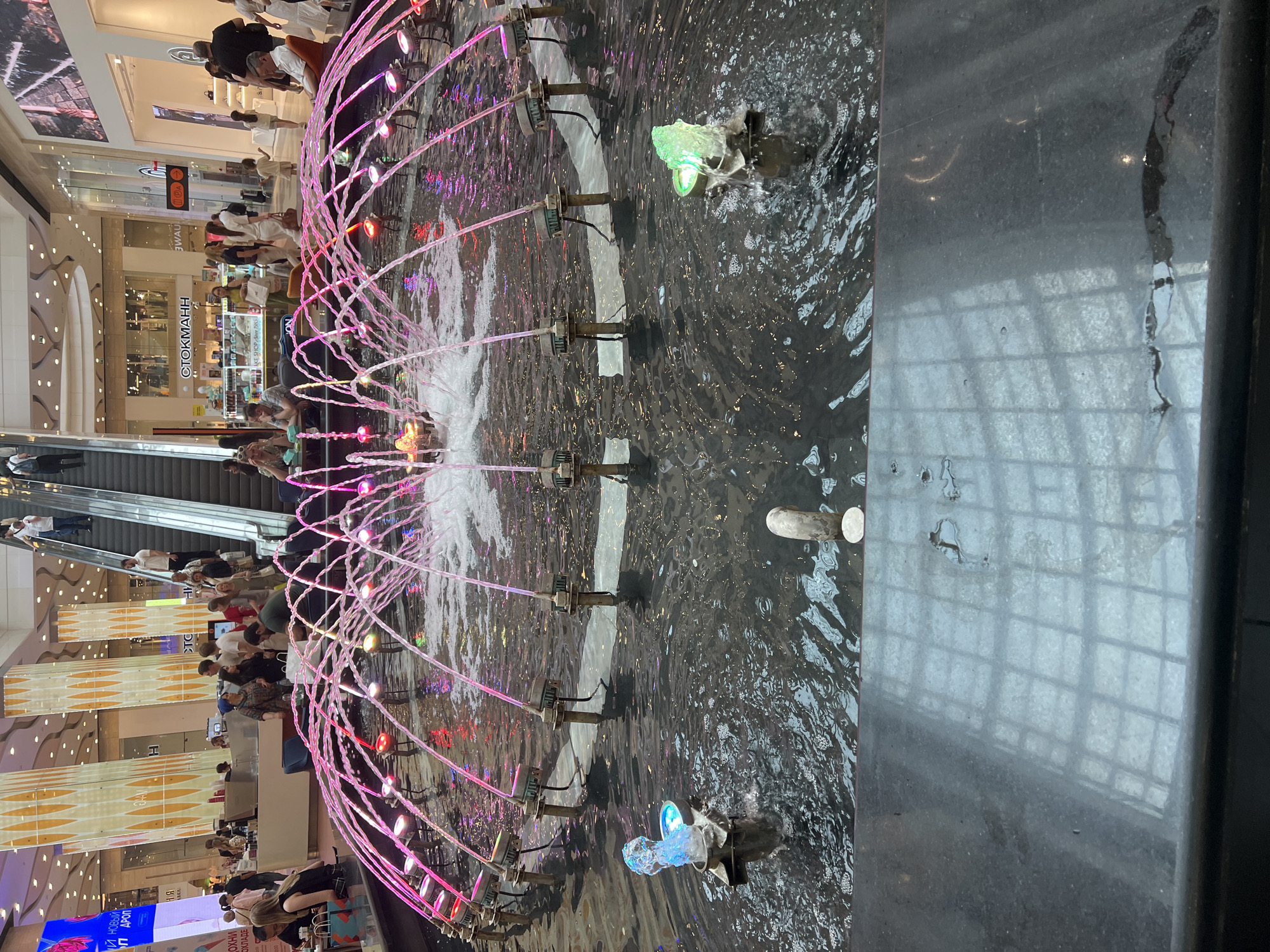 The image depicts a bustling indoor shopping mall, likely in Moscow, Russia, with a prominent water fountain in the foreground. The fountain features multiple jets of water illuminated by pink and purple lights, creating a vibrant and dynamic effect. Two smaller, individual water features, one emitting blue light and another green light, are visible to the left and right respectively.
In the background, shoppers are seen walking, browsing, and utilizing escalators. Various retail stores are identifiable, including "СТОКМАНН" (Stockmann), a well-known department store. The mall architecture is modern, with high ceilings, decorative pillars, and illuminated signage. The overall atmosphere is one of commercial activity and public gathering.