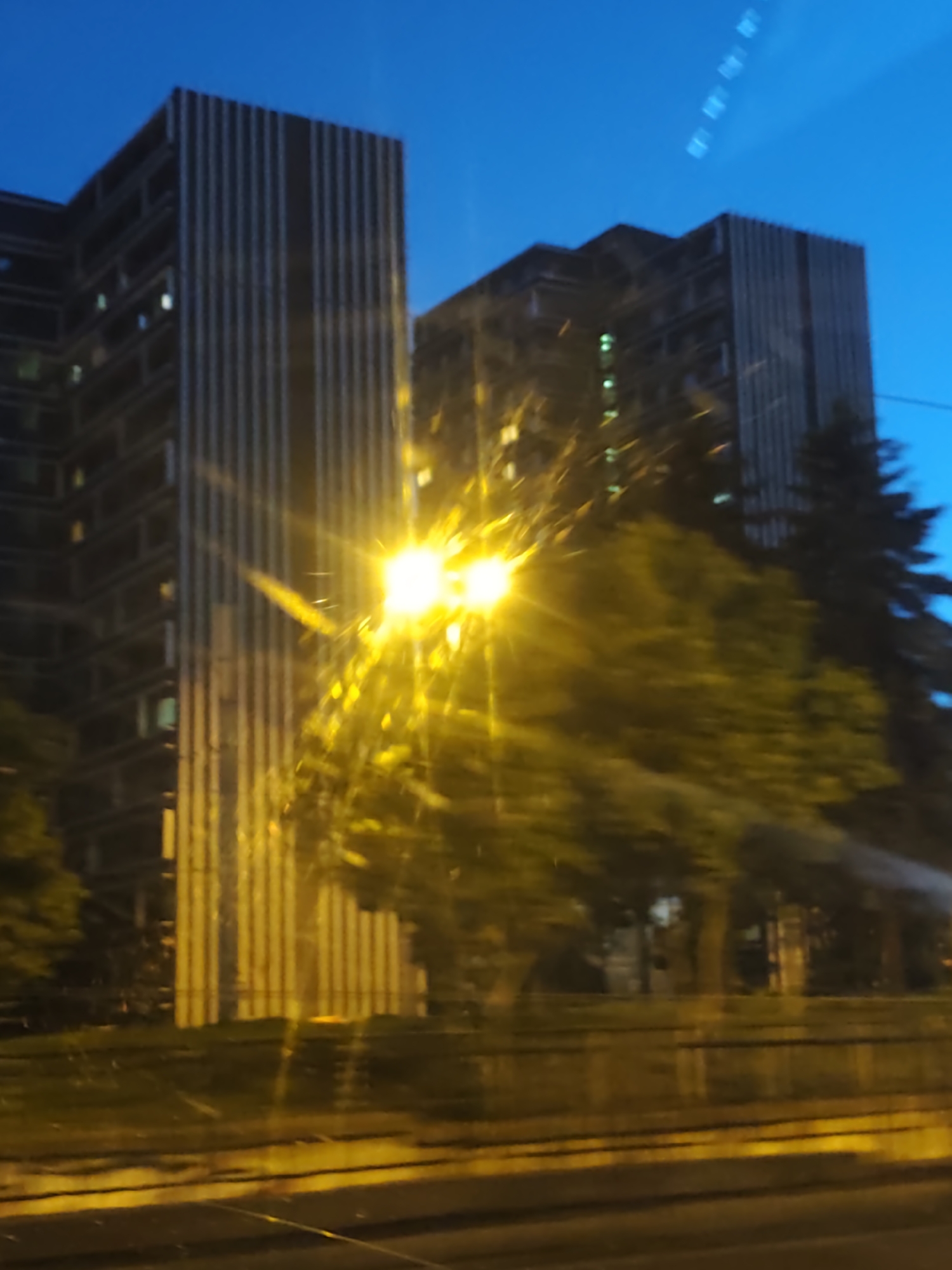The image depicts buildings and greenery, likely taken from a moving vehicle at night. Two tall buildings dominate the scene, their facades featuring vertical lines. One building appears closer and is partially illuminated by a bright yellow light, possibly from a street lamp. Trees and foliage are visible in the foreground, blurring due to the motion of the camera. The sky is a deep blue. Based on the architectural style of the buildings, it's possible that this photo was taken in Sofia, Bulgaria.