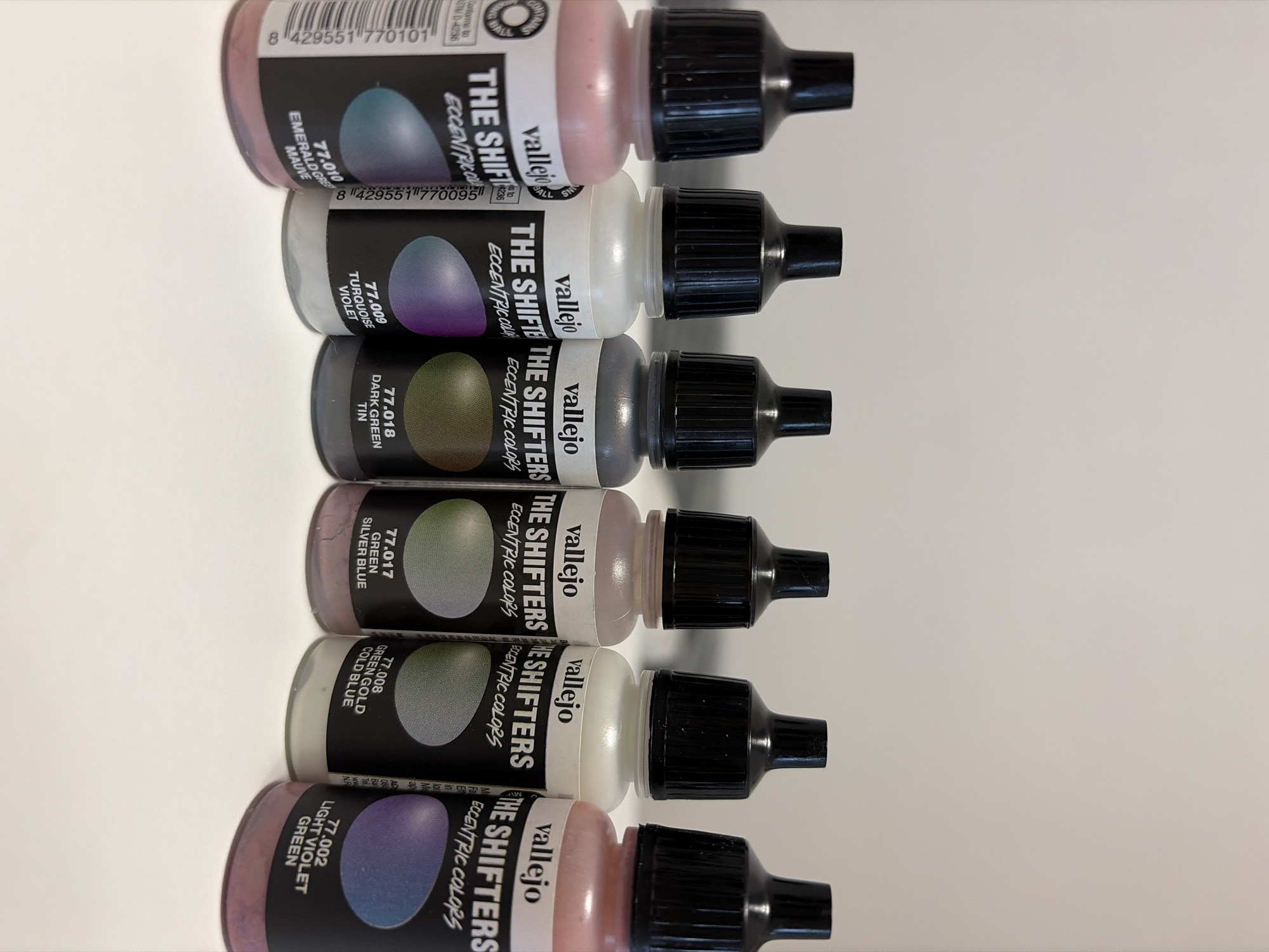 The image displays a stack of six paint bottles arranged vertically on a white surface. The bottles are all from the Vallejo "The Shifters" range, indicated by the prominent branding on each label. Each bottle has a black nozzle. The labels include a color-shifting effect, as implied by the name "The Shifters" and the phrase "Eccentric Colors."
From top to bottom, the bottle labels list the paint colors:
- 77.010 Emerald Green/Mauve
- 77.009 Turquoise/Violet
- 77.018 Dark Green/Tin
- 77.017 Green/Silver Blue
- 77.008 Green Gold/Cold Blue
- 77.002 Light Violet/Green
The image provides no information about location or people in Budva, Montenegro.