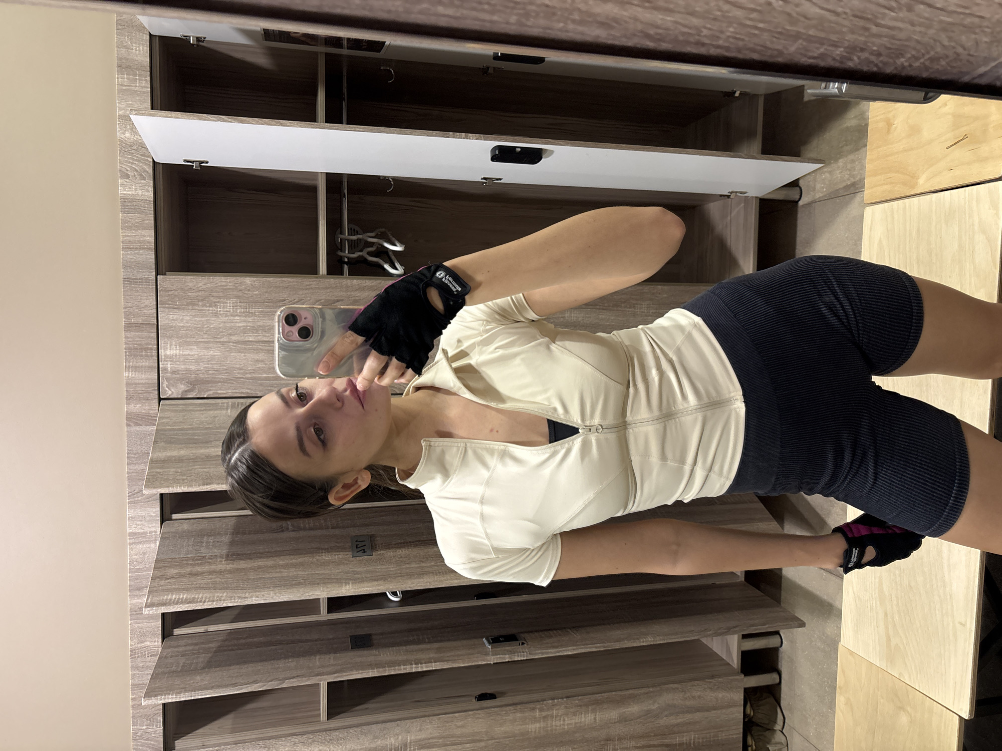 A young woman is taking a selfie in a locker room. She is wearing cycling shorts and a form-fitting shirt. The locker room appears to be in a gym or athletic facility. The background includes wooden lockers, some with open doors. The location is unknown, and no elements indicate Krasnoyarsk, Russia.
