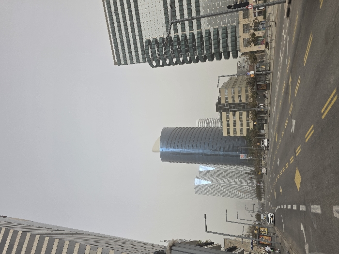 The image captures an urban street scene in Tel Aviv-Yafo, Israel, looking north along Begin Road towards the Azrieli Center area. The sky is uniformly overcast and hazy, presenting a light grey-brown hue.
The foreground consists of a multi-lane asphalt road marked with yellow double solid lines, dashed yellow lines, and white dashed lines. Yellow diamond-shaped markings and white directional arrows (straight, left turn, right turn) are painted on the road surface. Several vehicles are present: a white sedan or SUV is positioned in the left lane, and another white vehicle is further down the road in the right lane, with additional indistinct vehicles visible in the distance. A vertical traffic light pole is partially visible on the far right.
On the left side of the road, a sidewalk abuts a tall building with a striped facade, partially visible on the extreme left. Multiple grey, modern streetlights line the sidewalk. One individual is visible on a personal mobility device (e.g., electric unicycle or scooter) on the left sidewalk, accompanied by another pedestrian.
The midground and background are characterized by several high-rise structures. Centrally, the Azrieli Sarona Tower stands as a prominent, tall, cylindrical skyscraper with a reflective blue-tinted glass facade, crowned by a distinctive white, wing-like architectural feature. To its left are two other modern skyscrapers, identified as the Azrieli Town Towers, which exhibit reflective, geometrically patterned glass facades. Further right, a mid-rise building with a light-colored, multi-windowed facade is visible. On the far right, another tall building with a facade comprising dark horizontal bands and reflective surfaces partially frames the scene.
The right side of the street features a wide sidewalk equipped with multiple bus shelters displaying advertising panels, along with benches and additional streetlights. Indistinct figures of pedestrians are visible on this sidewalk. Sparse trees and bushes are present along the street edges.