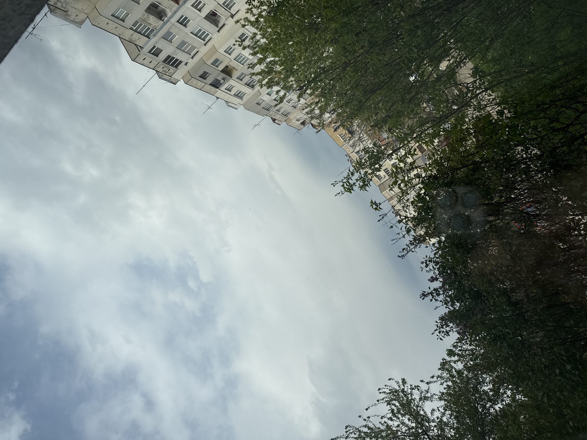 The image presents an upward-looking perspective of an urban environment, likely in Sofia, Bulgaria, characterized by architectural structures and natural elements under a cloudy sky.
The primary subjects are a multi-story residential building and dense green foliage. The building, visible across the top-left and top-right sections, is light-colored, possibly white or light grey, with numerous rectangular windows and several balconies. Some balconies appear enclosed or partially enclosed, while others are open. Small antennas or wires extend from the upper parts of the building. The architecture suggests a style common in Eastern European cities.
Framing the right and bottom-right sides of the image is a significant amount of vibrant green foliage from trees, indicating a spring or summer season. The dense leaves and branches partially obscure the view beyond them.
The central and lower-left portion of the frame is dominated by an overcast sky. The clouds are predominantly light grey and white, suggesting a cloudy or partly cloudy daytime, rather than direct sunlight or stormy weather.
No specific activity or event is taking place; the scene is static. There is no visible text. The overall impression is that of a typical residential area, observed from a low vantage point, possibly from a street or courtyard looking up between buildings. The image has a tilted orientation, making the buildings and trees appear diagonally aligned.