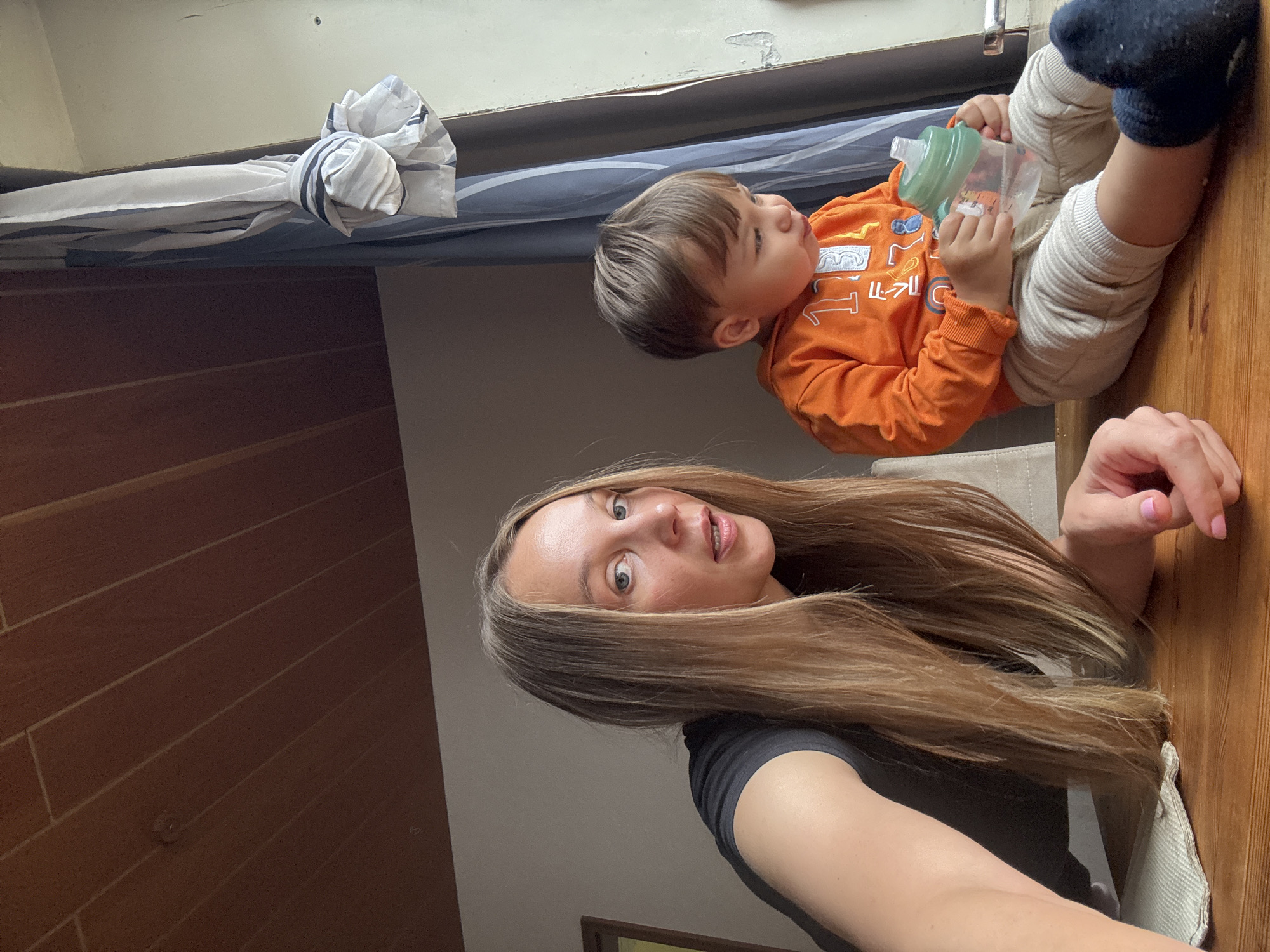 A woman with long blonde hair and a black t-shirt is leaning forward on a wooden table, looking at the camera. Next to her, a toddler in an orange sweatshirt and beige pants is holding a sippy cup. The toddler is looking down at the cup. The scene appears to be indoors, with a wooden ceiling above and a window with curtains to the right. The overall impression is a casual, domestic setting.