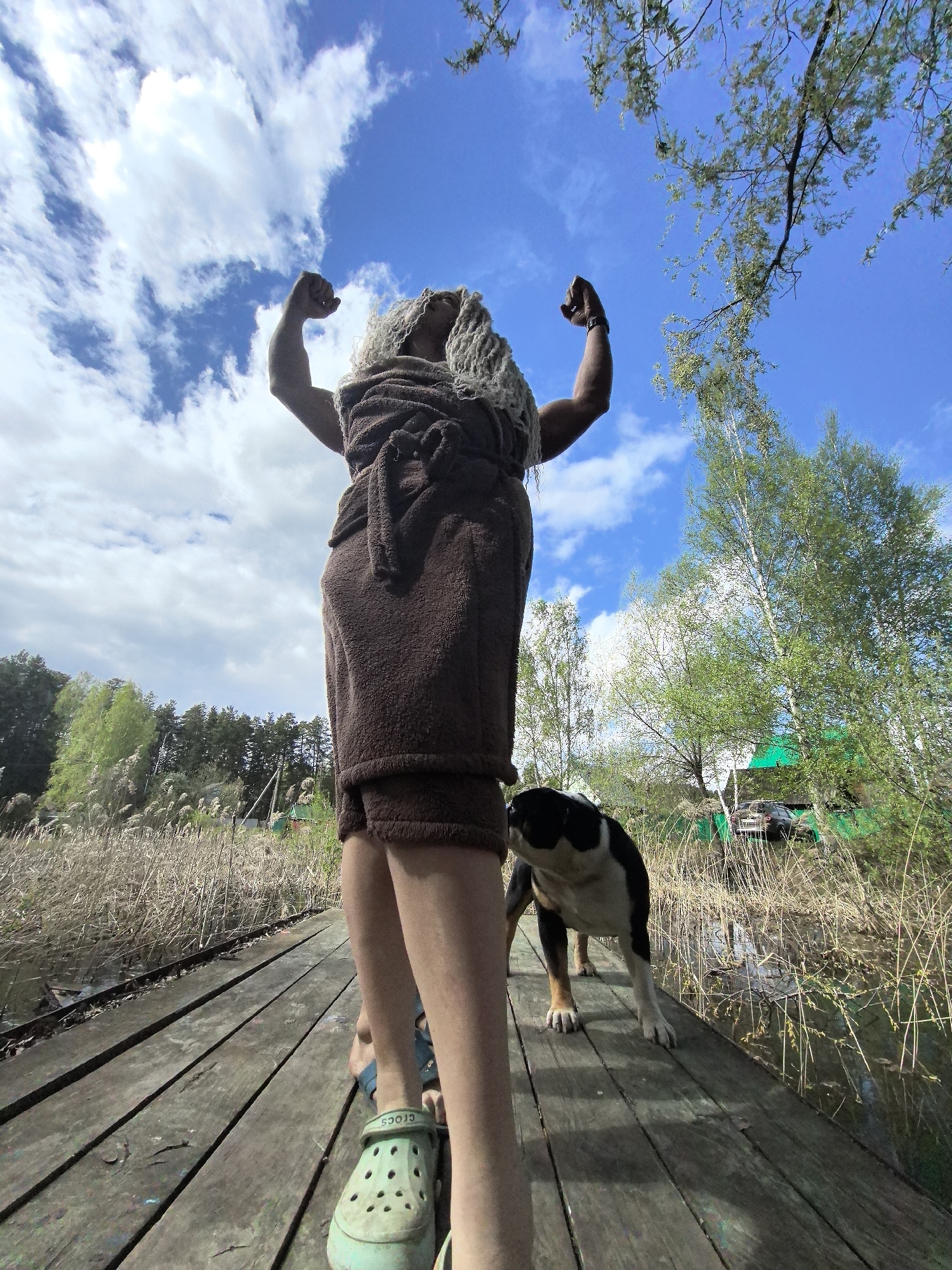 The image depicts a person standing on a wooden pier, flexing their arms. They are wearing a brown towel-like garment and green Crocs. The person has what appears to be a blonde wig on their head. To the right of the person, a black and white dog stands on the pier, looking towards the reeds and water surrounding the dock. The pier is situated in a swampy area with tall reeds visible in the background. Beyond the reeds, there is a green-fenced area with a vehicle parked inside. The sky is blue with white, puffy clouds. The location is identified as Noviy, Russia.
