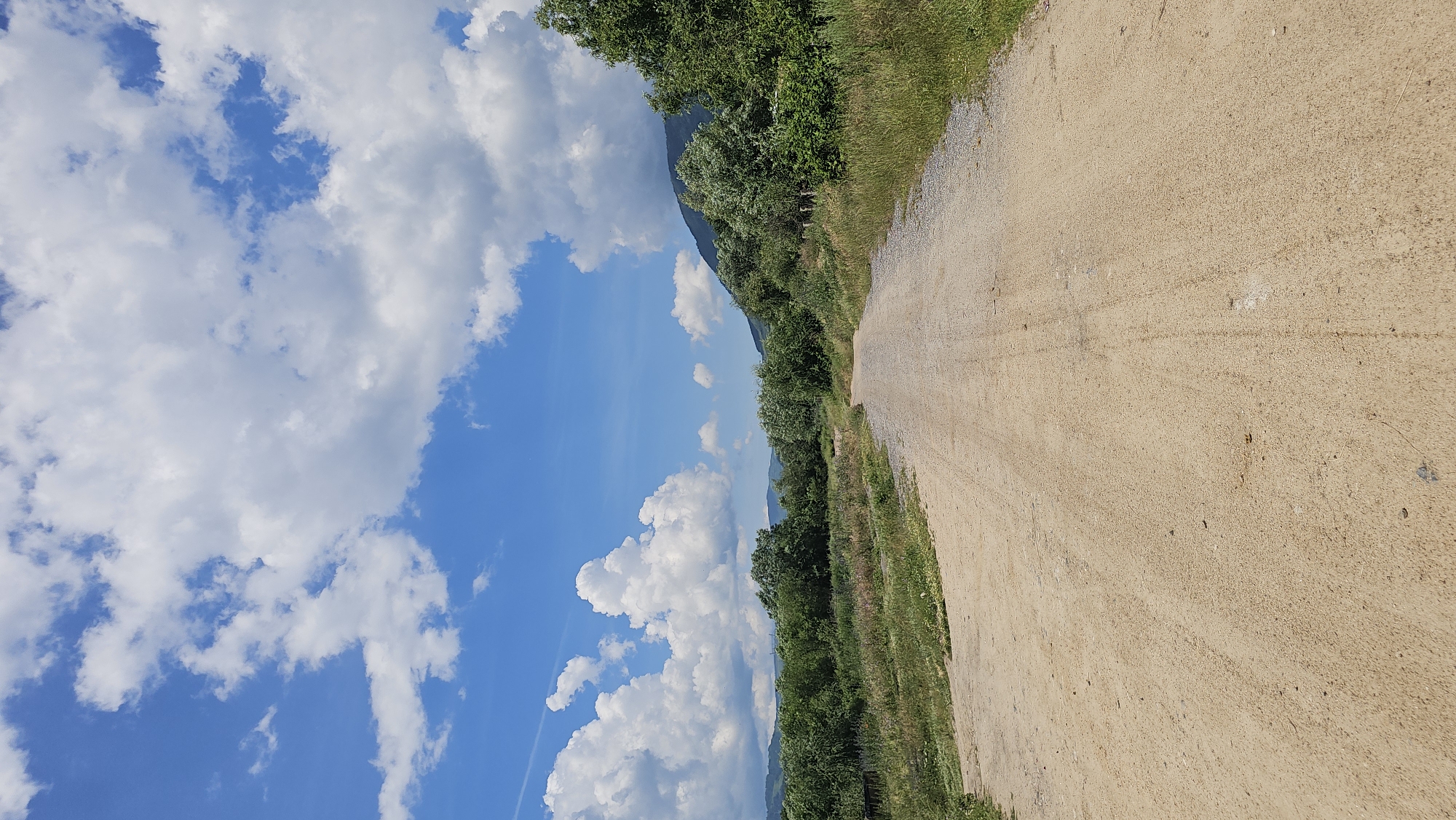 A dirt road curves through a rural landscape under a bright blue sky dotted with fluffy white clouds. The road, composed of light brown dirt and gravel, stretches into the distance, flanked by green vegetation, trees, and rolling hills. The scene appears to be a daytime view in the village of Rozino, Bulgaria.