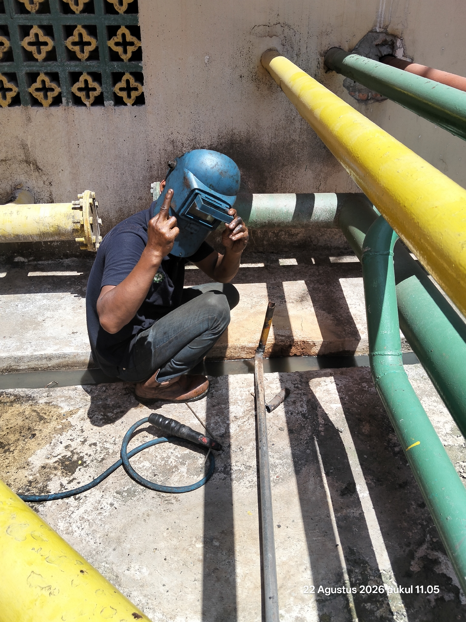 A person wearing a blue welding helmet and dark clothing is kneeling on a concrete surface outdoors, holding their left hand up to the helmet and their right hand in a fist. A welding torch with a blue cable is on the ground to the left. In the background, there are several large pipes, one yellow and several green, running horizontally and vertically. A section of a wall with a decorative pattern is visible in the upper left. The ground is a weathered concrete surface with shadows cast by the pipes. A date and time stamp, "22 Agustus 2026 pukul 11.05", is present in the lower right corner.