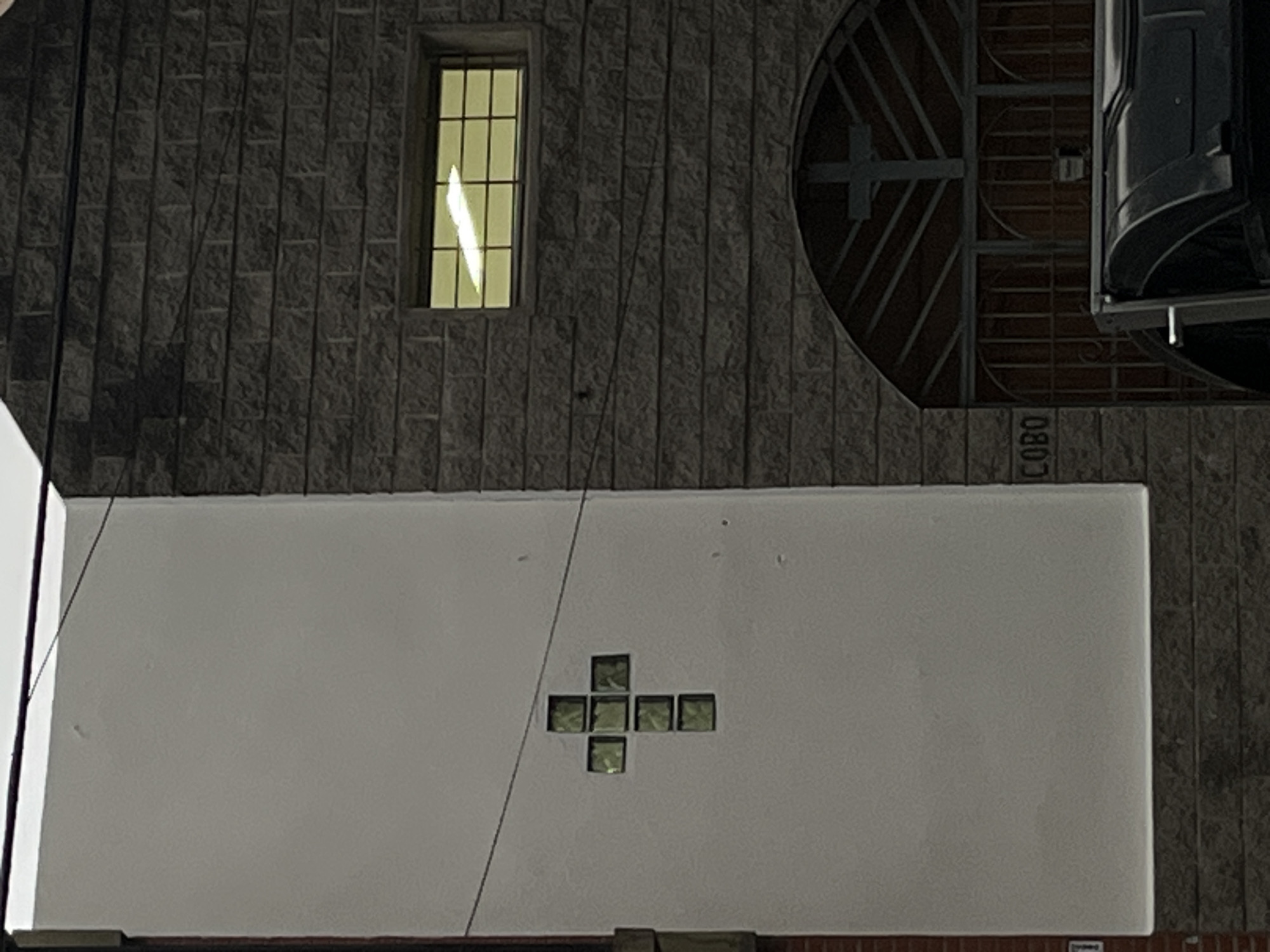 The image depicts a section of a building facade. The building has a combination of stone and white plastered walls. A single rectangular window is visible on the upper stone section, with its interior lit. Adjacent to it is a circular window with a cross-like design. Underneath, a white plastered wall has a cross-shaped pattern made of smaller translucent squares. The word "LOBO" is inscribed vertically on the stone wall. A portion of a vehicle is visible near the circular window. The image seems to be taken at night.