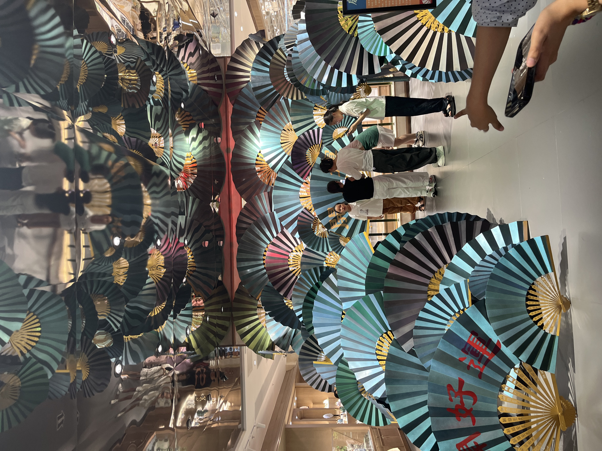 In the Haizhu District of China, a group of people are gathered in what appears to be a commercial setting, possibly a mall or a department store. The environment is decorated with a large display of fans, both in the foreground and reflected in the mirrored ceiling above. The fans are primarily in shades of teal, black, and gold, with some red Chinese characters visible on a few of them in the foreground. The characters on one fan appear to read "八好运" (bā hǎo yùn), which translates to "Great Luck."
In the mid-ground, four individuals are standing and interacting. One person, wearing a white t-shirt and black pants, is looking at a phone. To their left, another person in a white t-shirt and white pants is also engaged with the display. Further left, a person in a white t-shirt and brown pants stands looking in the same direction. To the right of the person on the phone, another individual in a white t-shirt and black pants is present. The individuals appear to be customers browsing the display.
In the foreground, a person's hand is visible reaching down towards a black object, possibly a phone or a small tray. This person is also making a gesture with their thumb down. The floor is a light grey tiled surface. In the background, beyond the fan display, shelves and displays of merchandise are visible, suggesting a retail environment. The lighting in the space is bright, with spotlights visible in the ceiling reflection.