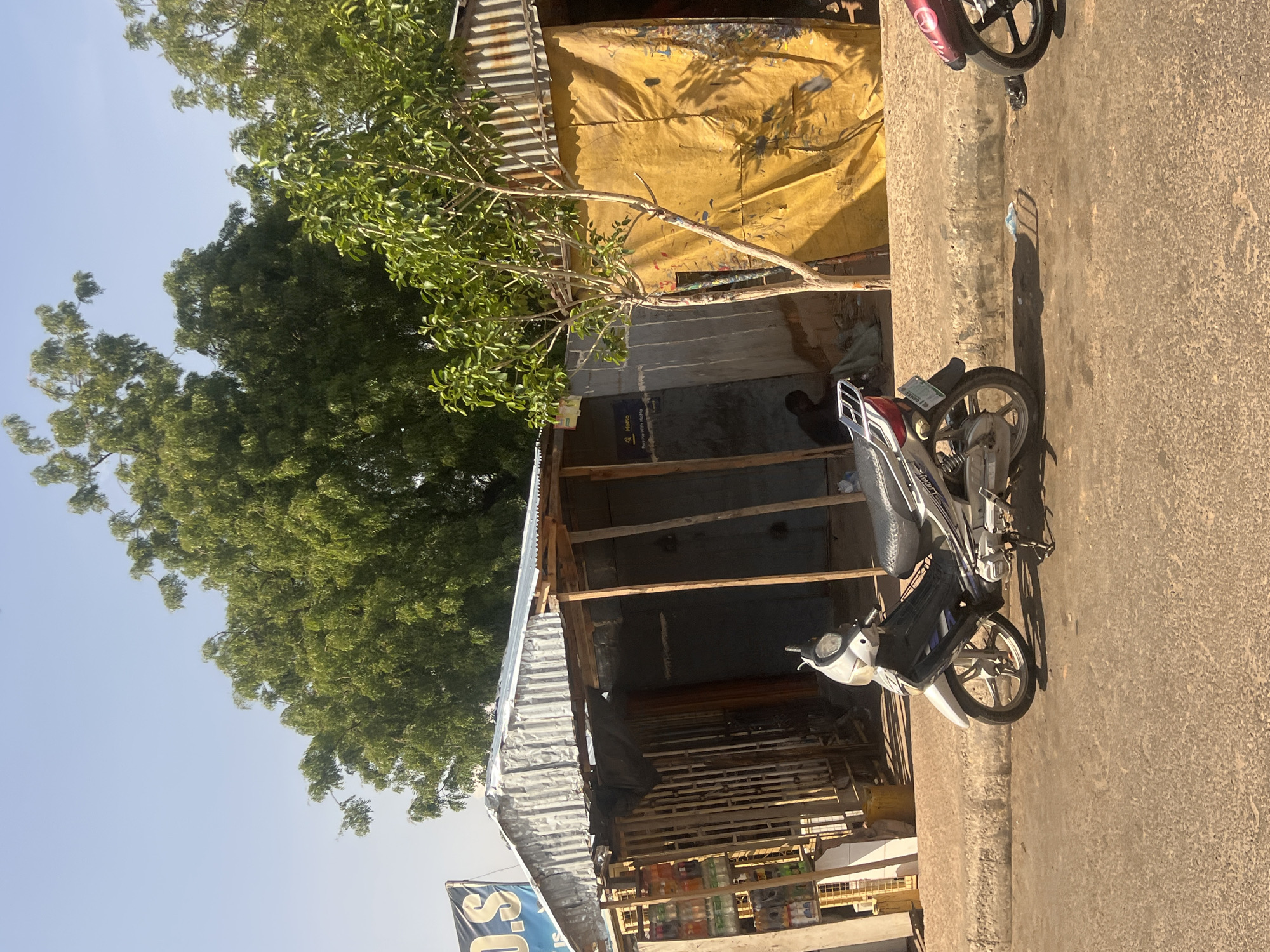 The image captures a sunlit street scene in Gombe, Nigeria, characterized by roadside stalls and parked motorcycles. In the foreground, an asphalt road with visible texture is separated from a dirt and gravel shoulder by a concrete curb. Three informal structures line the shoulder. The leftmost stall features a corrugated metal roof, wooden support beams, and open shelving stocked with various containers, possibly beverages; a partial blue sign reading "O.S" or "D.S" is affixed above. Adjacent to this, a second open-fronted structure, also with a corrugated roof, contains a person seated or crouching within its dark interior. To the right, a third structure under a corrugated metal roof is largely enveloped by a stained yellow tarp, with a small, leafy tree positioned directly in front of it. A "MoMo Pay Me With MoMo" sign is visible on the wall behind this tree. Two motorcycles are present on the road: a white and silver "LUCKY" model is parked centrally on its kickstand, facing left, while the front wheel and fender of a darker-colored motorcycle are partially visible on the far right. A dense canopy of large green trees dominates the background against a clear, pale blue sky. The scene is illuminated by bright sunlight, generating distinct shadows.