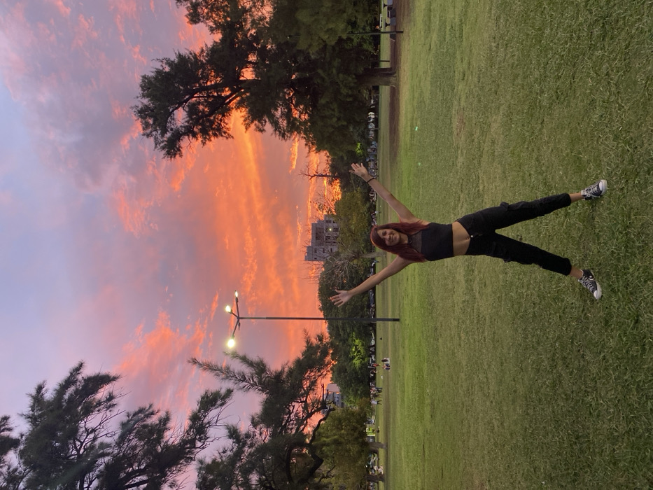 A young woman in a black crop top and pants poses with arms outstretched on a grassy field. Behind her, a vibrant sunset paints the sky in shades of orange, pink, and purple. Trees frame the scene, and a lamppost stands to the left. The buildings in the distance and the people in the center ground indicate that this shot was taken in the late afternoon, probably in a park in an urban area.