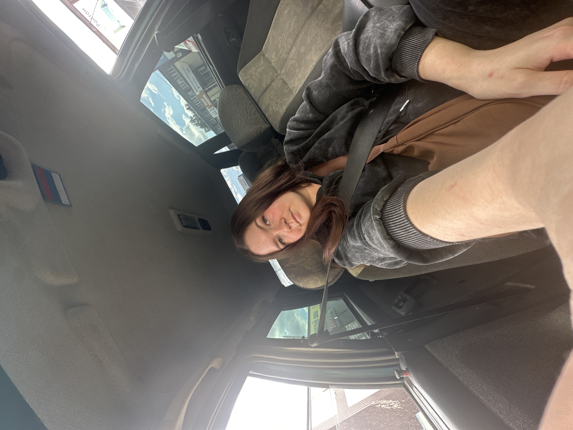 A young woman with long brown hair sits in the passenger seat of a car, looking directly at the camera. She is wearing a dark grey, mottled hoodie and a brown shoulder bag is visible. A seatbelt is fastened across her. The interior of the car is visible, including the headrest, car roof with a small Russian flag sticker, and the window. Through the window, buildings and trees can be seen, suggesting an urban or suburban environment. A sign with Cyrillic lettering is visible, with the text "РОЙТОН" and "ОТКРЫТИЕ". Based on the text on the sign, the location is likely Chaikovsky, Russia. The sky is blue with some clouds. The image appears to be a selfie taken from the driver's perspective.