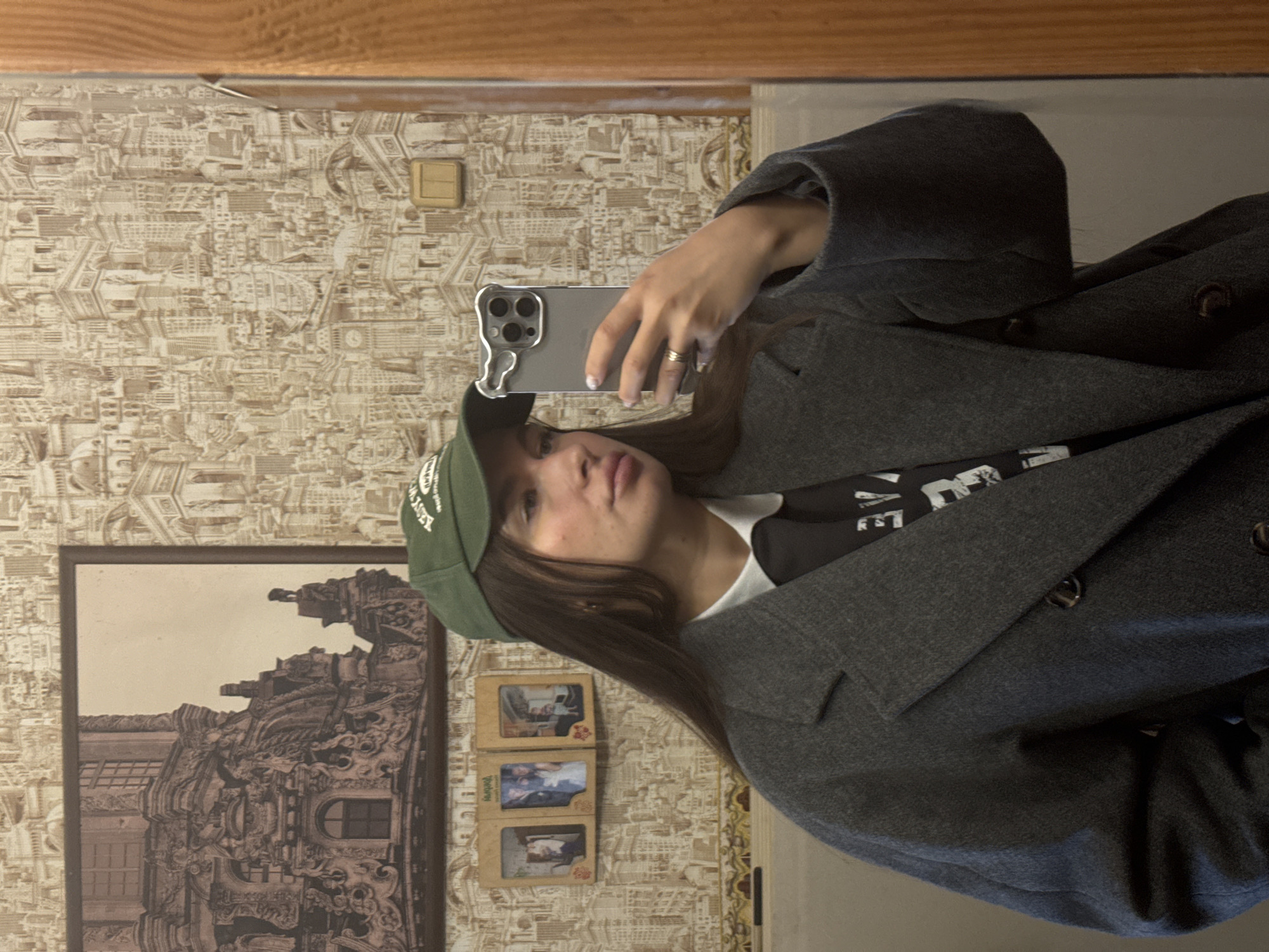 The image shows a woman taking a selfie in a mirror. She is wearing a green baseball cap, a black t-shirt with white lettering, and a gray coat. She is holding a smartphone in her right hand.
Behind her, the wallpaper has a repeating pattern of cityscapes. There are several framed pictures on the wall including a large picture of architecture and several small pictures of people. There is a light switch on the wall.
The location cannot be determined to be in Красный Маяк, Russia, based on the visual information provided.