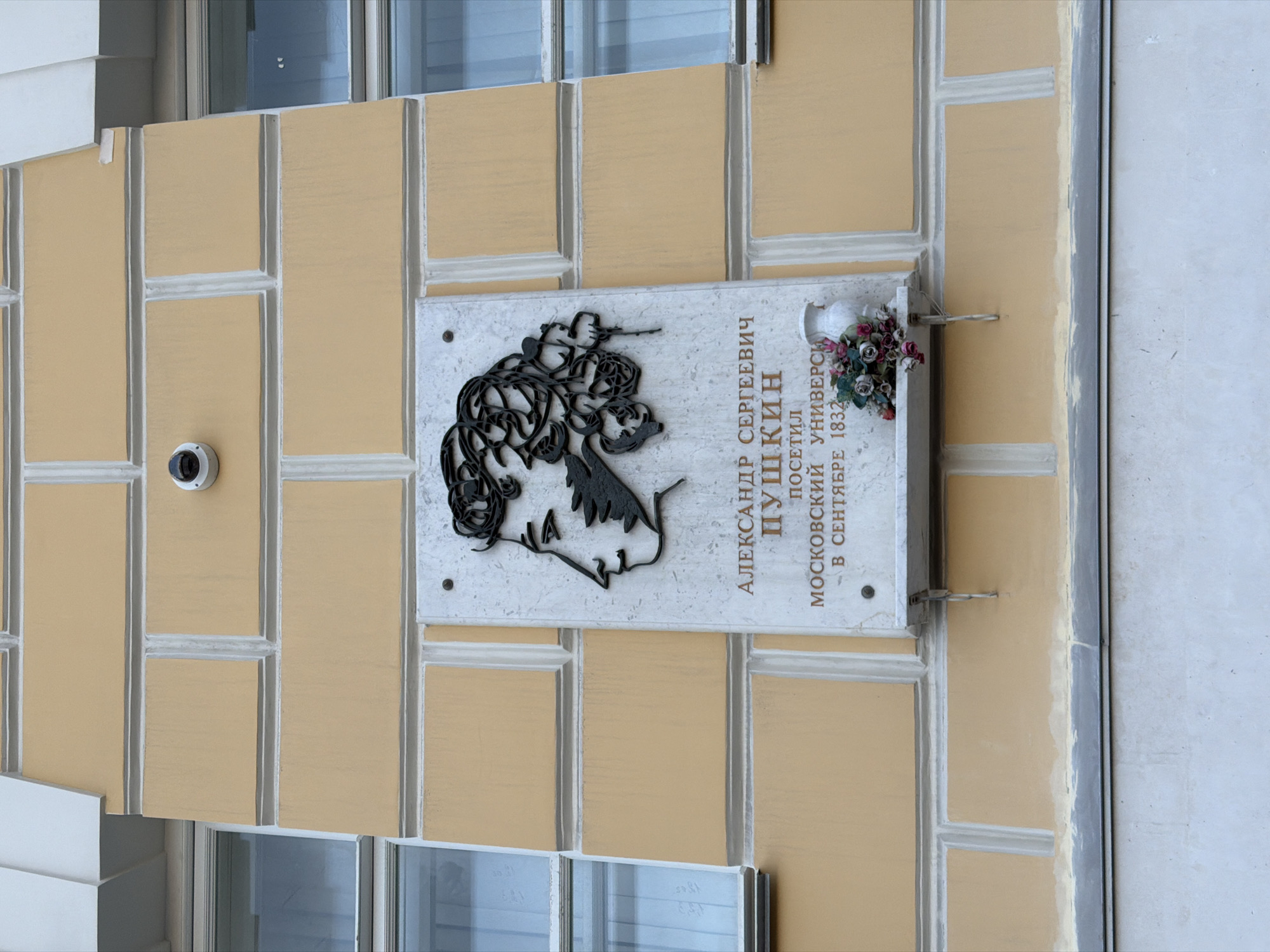 The image shows a plaque on a building in Moscow, Russia. The plaque is dedicated to Alexander Pushkin and indicates that he visited Moscow University in September 1832. The plaque features a black silhouette of Pushkin's profile and is adorned with a small vase of flowers. A security camera is mounted on the wall to the left of the plaque. The building is a light yellow color, with a grid-like pattern on its façade. A window is visible below the plaque. The image is taken from a slightly low angle, and the plaque is positioned on the right side of the frame.