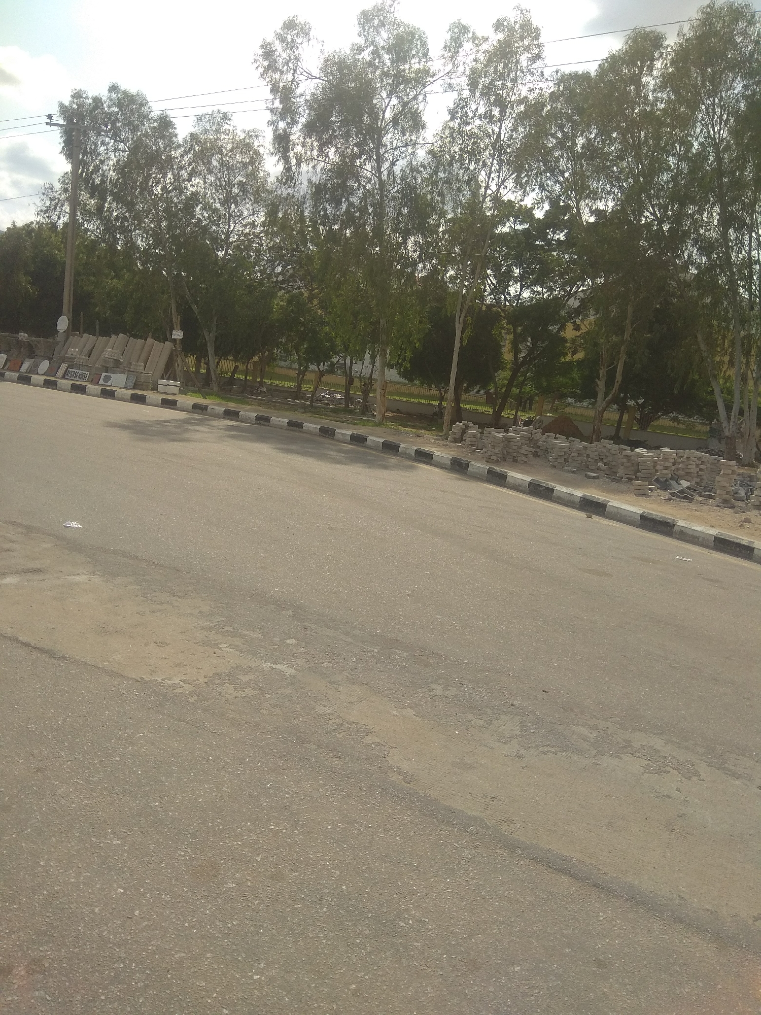 The image shows a street scene in Jalingo, Nigeria. A paved road occupies the foreground, with a sandy shoulder to the left. Along the shoulder are stacks of paving stones and other construction materials, suggesting ongoing or recent roadwork. A decorative curb with alternating black and white sections runs along the edge of the road. Beyond the curb, a line of tall trees with thin trunks and dense foliage forms a green barrier. Power lines are visible in the sky above the trees. The overall impression is that of a semi-urban environment with clear signs of infrastructure development.