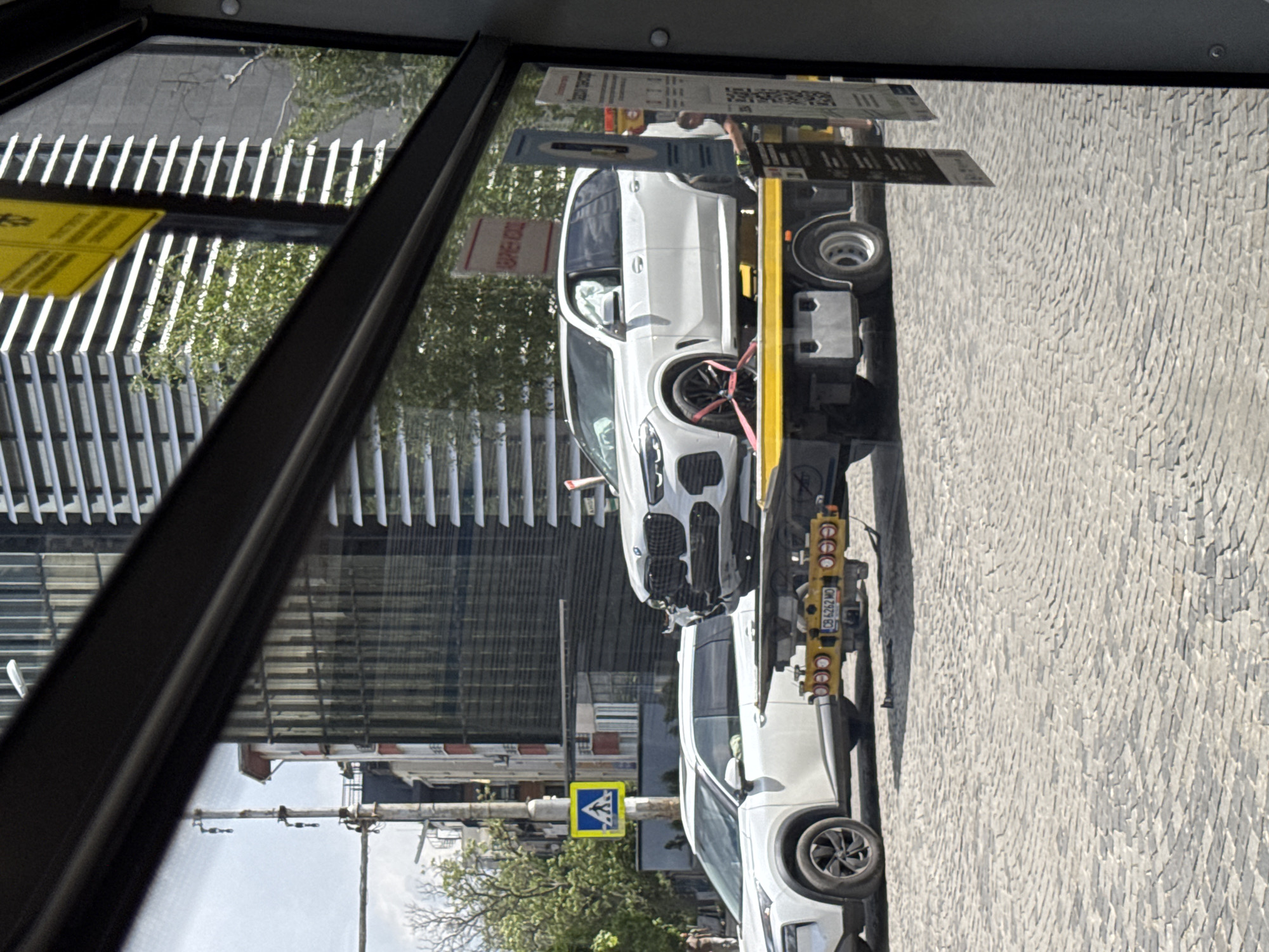 From inside a bus or tram in Sofia, Bulgaria, a view through a dirty window shows a white damaged BMW X5 being towed. The damage to the front of the vehicle appears consistent with a car accident. The X5 is secured with red straps to a yellow tow truck. The street is cobblestone. Reflected in the window is a modern building with louvered sunshades. A pedestrian crossing sign is visible in the background.