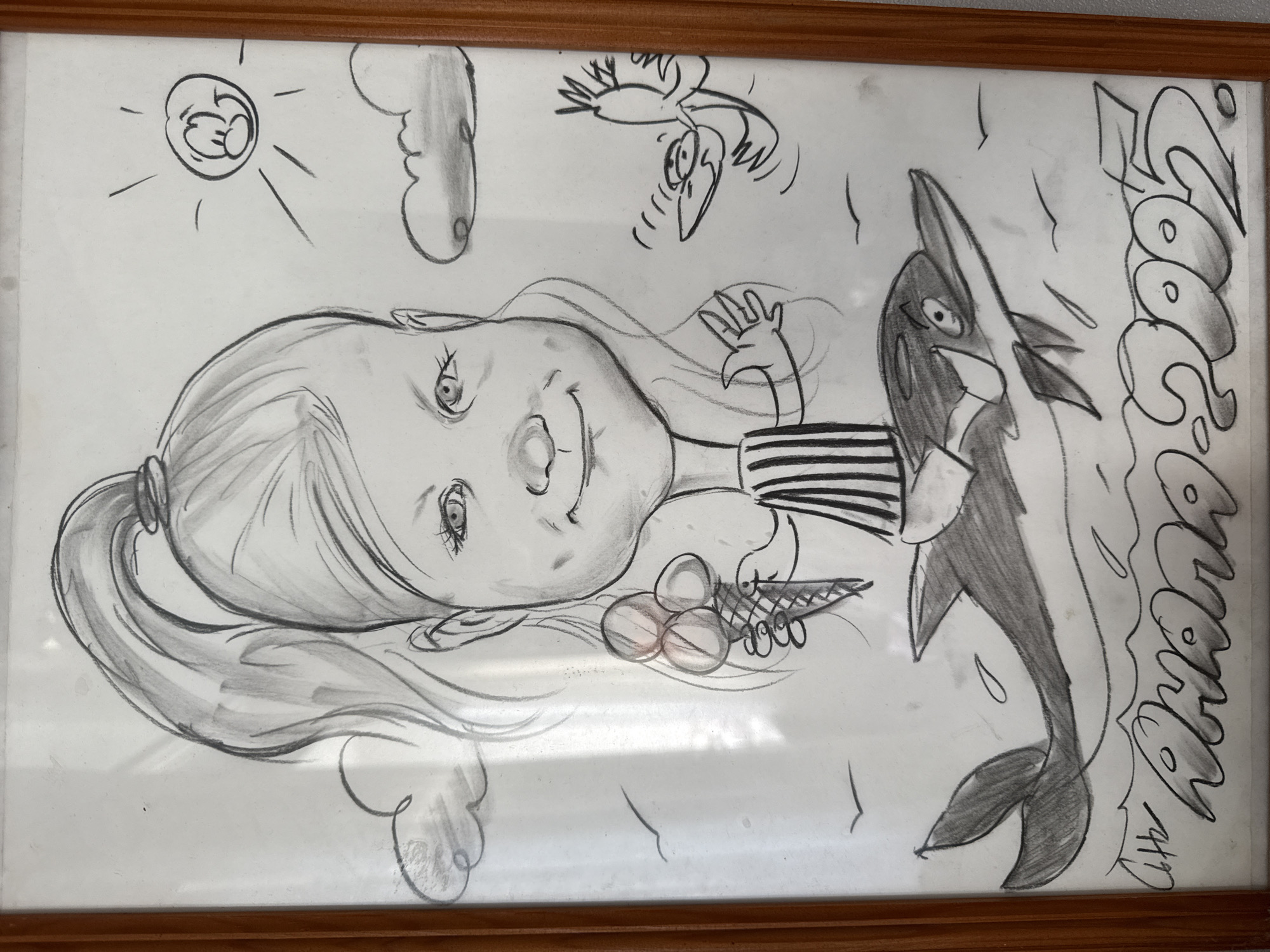 A framed drawing depicts a stylized scene. A girl with a large head, light hair in a ponytail, and a striped shirt holds a triple-scoop ice cream cone. A cartoonish orca is beside her, and a bird flies above. A sun with a cartoon face and a cloud are in the upper left, while a cloud is below the girl. The word "Chaika" is written vertically along the right edge. A signature with the date "4/17" is in the bottom right corner. The drawing is in a brown wooden frame. This image likely depicts an event or memory from the city of Chaikovsky, Russia.