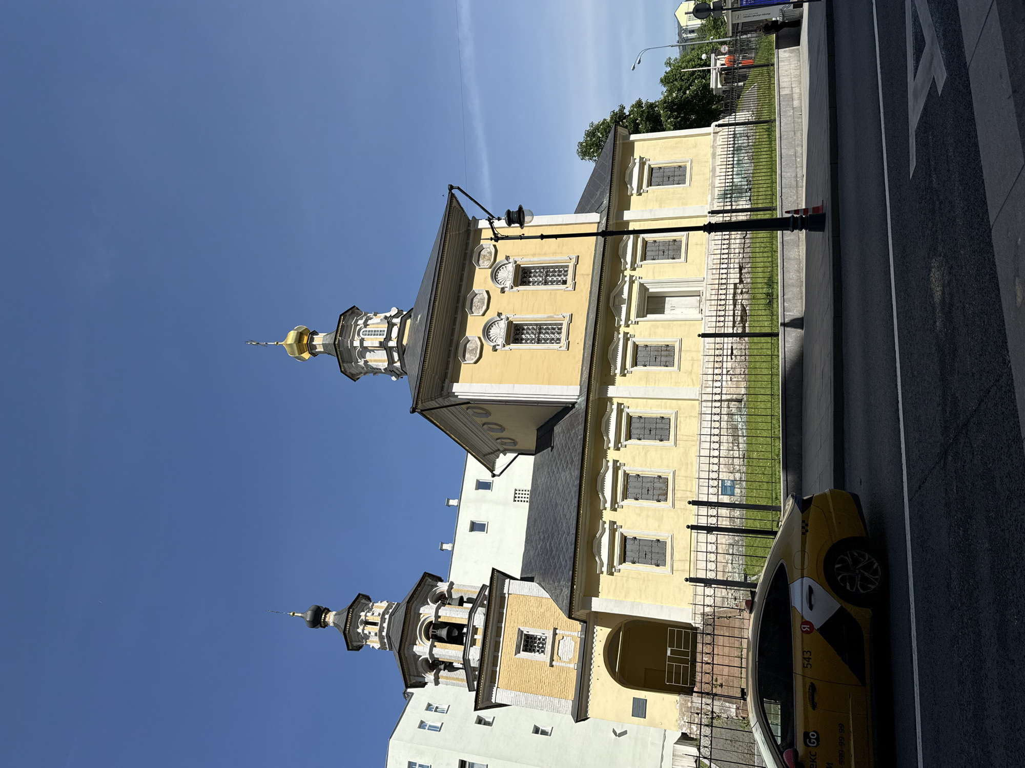 The image depicts a yellow building with a steeple in Moscow, Russia. The building has multiple windows and decorative elements. A black metal fence surrounds the building, with grass visible inside the fenced area. A street and a yellow taxi are present in the foreground. The sky is blue and clear.