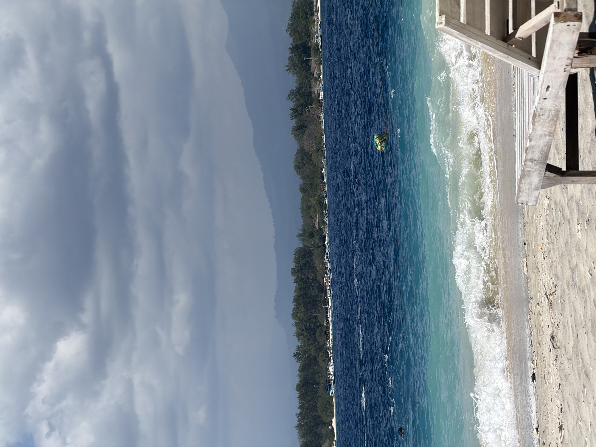 A vertical shot captures a serene beach scene in an unknown Indonesian location under a cloudy sky. The foreground features a weathered wooden lounge chair on a sandy beach, with small stones scattered across the sand. Gentle waves with foamy crests roll onto the shore, transitioning from a turquoise hue to a deeper blue as they recede. In the mid-ground, a lush green coastline is visible, lined with trees and dotted with small buildings and numerous boats anchored near the shore. Beyond the coastline, hazy mountains form the backdrop. A yellow and green buoy floats in the water, connected to a rope. The overall atmosphere is peaceful and tranquil, with the natural beauty of the landscape taking center stage.