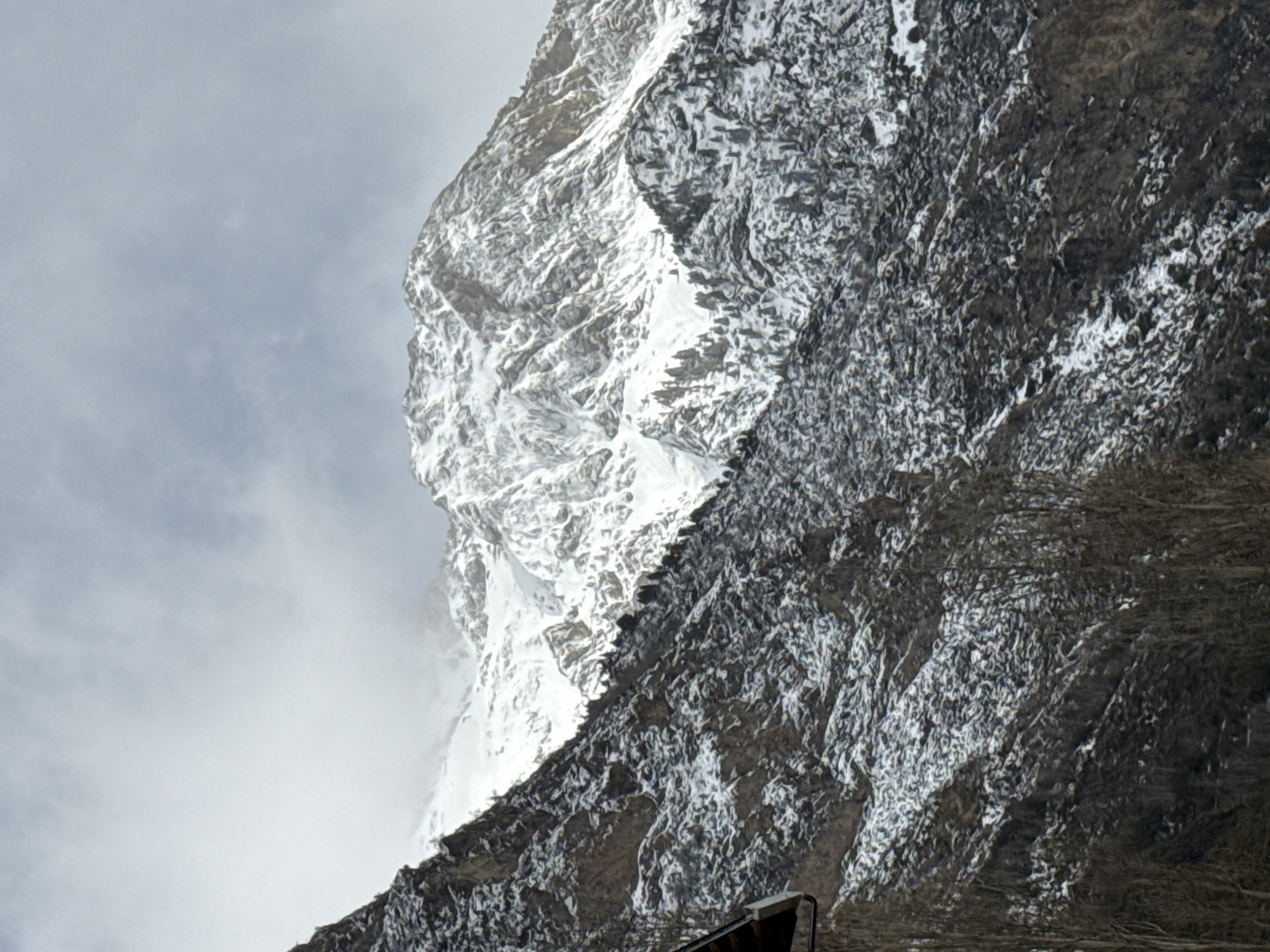 A ground-level, medium shot shows a high-elevation mountain face. The left face is heavily covered in ice and snow and lit by sunlight. The right face of the mountain is bare rock with snow and ice mixed into the crags. The mountain is set against a gray sky and fog to the left. A small human-made structure is at the bottom right of the frame. This is likely a mountain in the Pyrenees in Benasque, Spain.