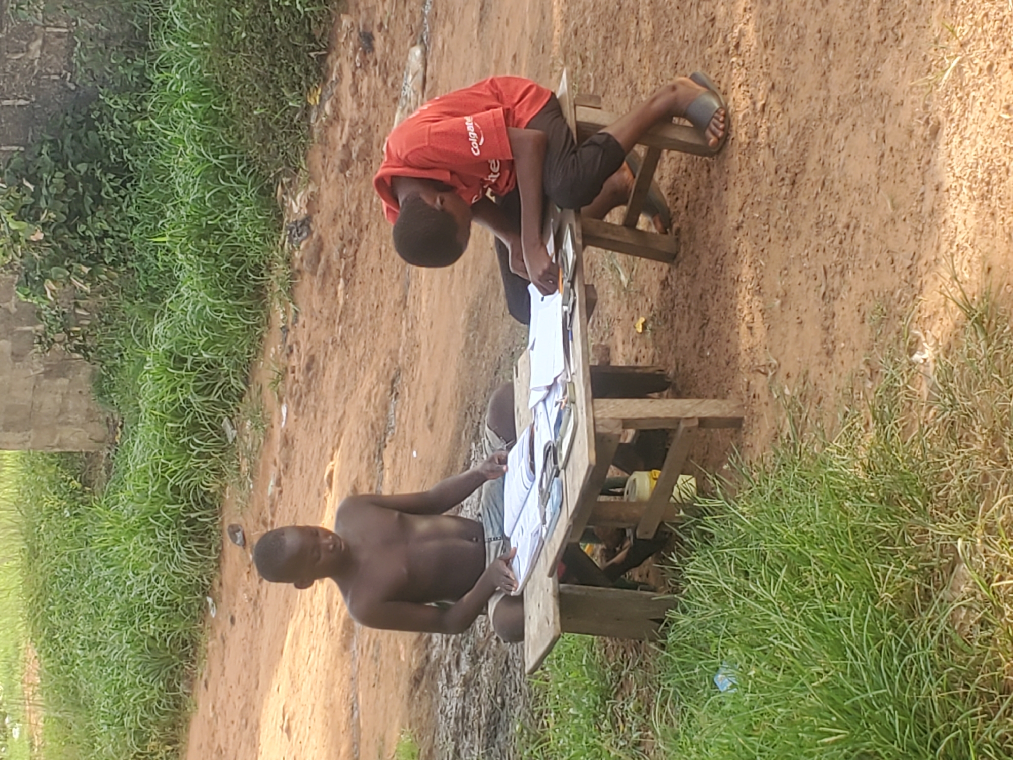 Two young boys are seated at a wooden desk in Ukwu-Abwa, Nigeria. One boy is wearing a red "Colgate" t-shirt and dark shorts. He is bent over the desk, appearing to be working on something. The second boy is shirtless and wearing light-colored shorts. He is seated and seems to be interacting with papers on the desk. The desk is constructed of wood and set up on a dirt ground outdoors with green grass and vegetation in the surrounding area. A building is visible in the background.