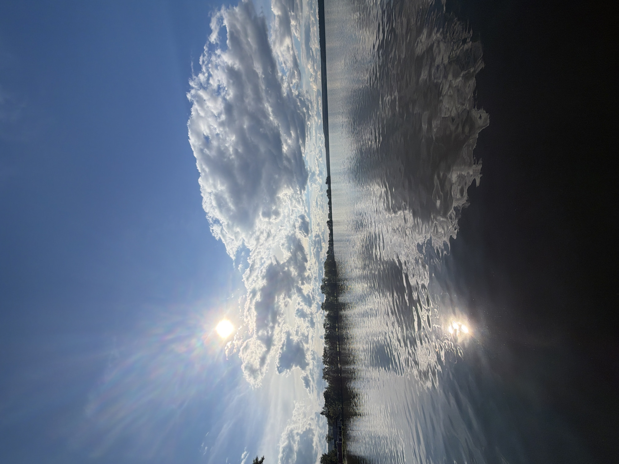 The image shows a view of a lake in Votkinsk, Russia, under a bright blue sky with scattered white cumulus clouds. The sun is visible, creating a starburst effect. The lake's surface reflects the sky and clouds, creating a near symmetrical composition. The shoreline is visible in the distance, with trees lining the edge of the water. The reflection of the sun in the water is also visible.
