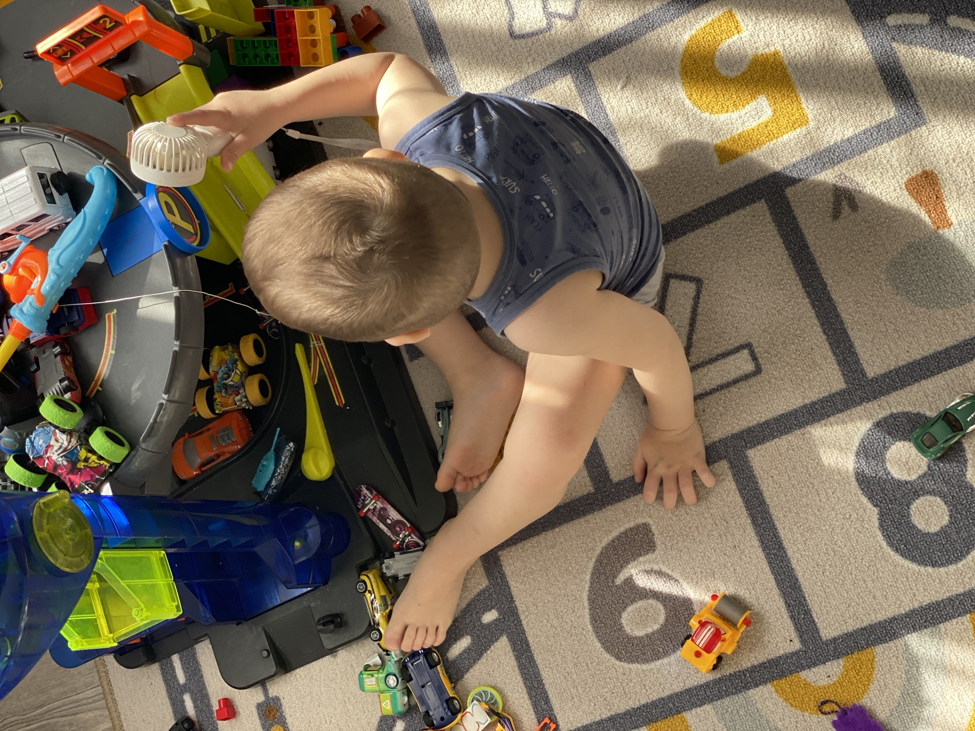 A young child with short brown hair is sitting on a number-themed rug, surrounded by toys. The child is wearing a navy blue shirt with white patterns. The rug displays numbers in yellow and gray. Toys include various cars, trucks, building blocks, and a racetrack. The setting is indoors and there is no clear indication of it being located in Sochi, Russia.