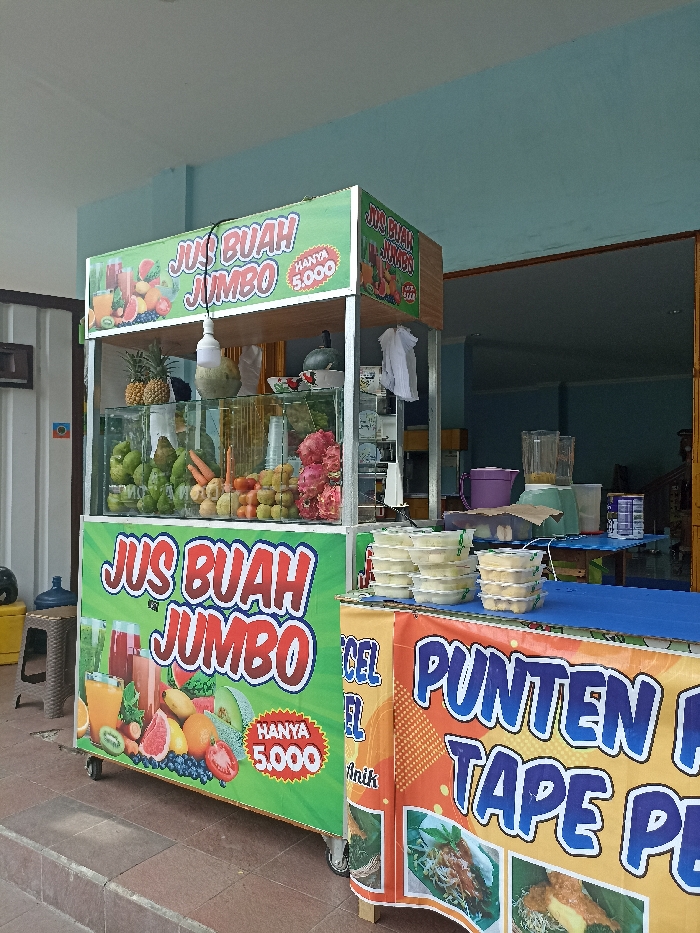 This eye-level shot captures two mobile street food stalls set up outdoors in Tulungagung, Indonesia, against a light blue building. The bright, clear lighting suggests it is daytime.
The prominent stall on the left is a "Jus Buah Jumbo" (Jumbo Fruit Juice) stand, featuring vibrant green and white signage. The banners display images of various fruits and juices, along with the text "JUS BUAH JUMBO" and "HANYA 5.000" (Only 5,000), indicating the price in Indonesian Rupiah. A transparent display case on the stall's counter holds an assortment of fresh fruits, including pineapples, guavas, carrots, tomatoes, grapes, and dragon fruits. A hanging light bulb illuminates the display. To the left of this stall, a blue water dispenser and a small stool are visible.
Adjacent to the juice stall, on the right, is another food stall with an orange and blue banner. Partial text on this banner reads "PUNTEN" and "TAPE PL," likely advertising traditional Indonesian snacks like Punten and Tape. Small images of food dishes are also depicted on the banner, along with the word "Anik" and partially "CEL". The stall's blue tabletop is stacked with several clear plastic containers, possibly holding prepared portions of food. In the background behind this stall, blenders and other kitchen equipment are visible, suggesting a food preparation area. No people are visible in the frame, but the setup indicates an active vending scene.