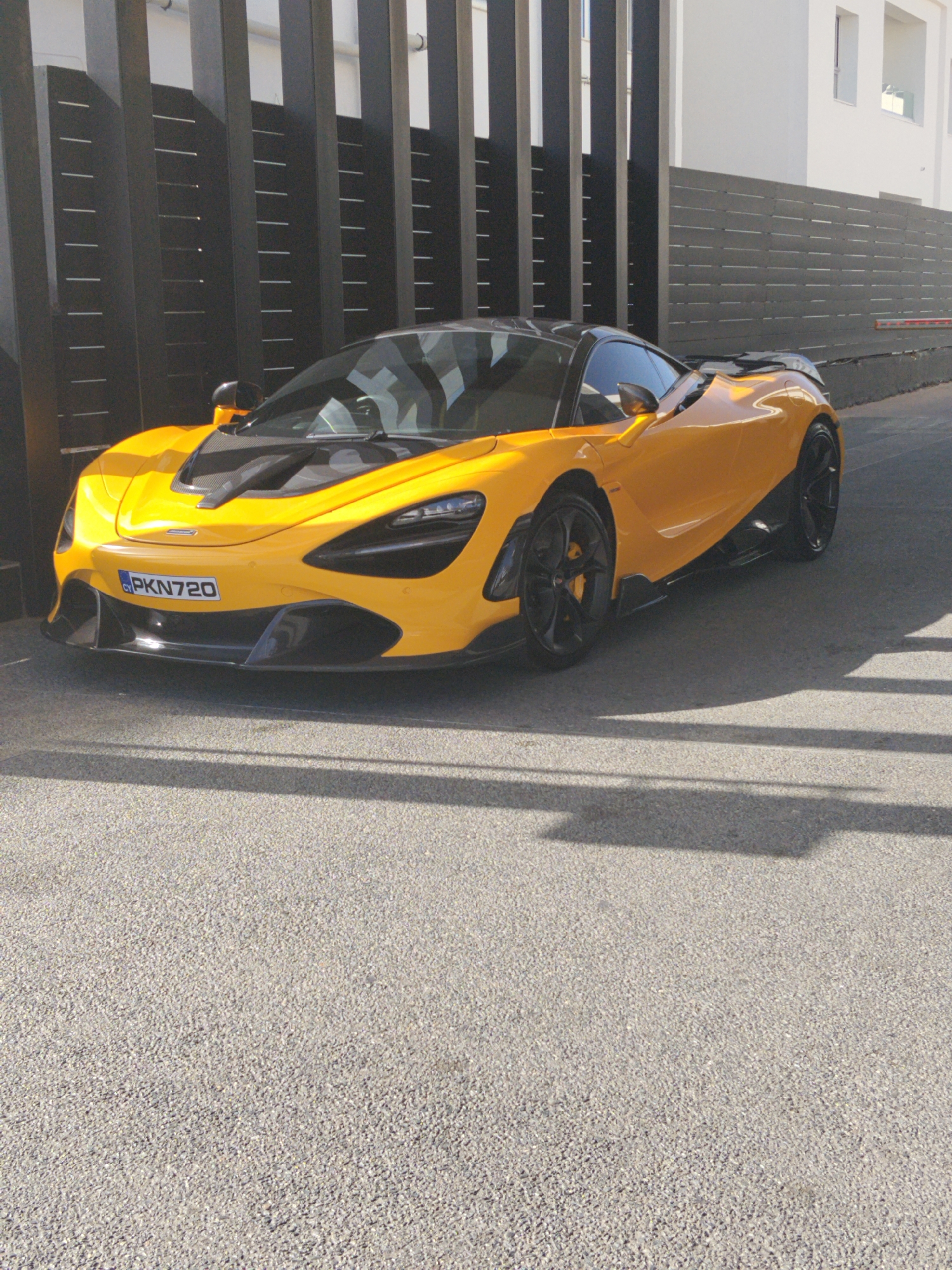 A McLaren 720S sports car, painted yellow with black accents, is parked on a gray asphalt surface. The license plate reads "PKN720." The car is situated in front of a modern metal fence composed of vertical bars with horizontal slats. Behind the fence is a white building. The location is Ayia Napa, Cyprus. The scene is bathed in sunlight, casting shadows across the asphalt. There are no people visible in the image.