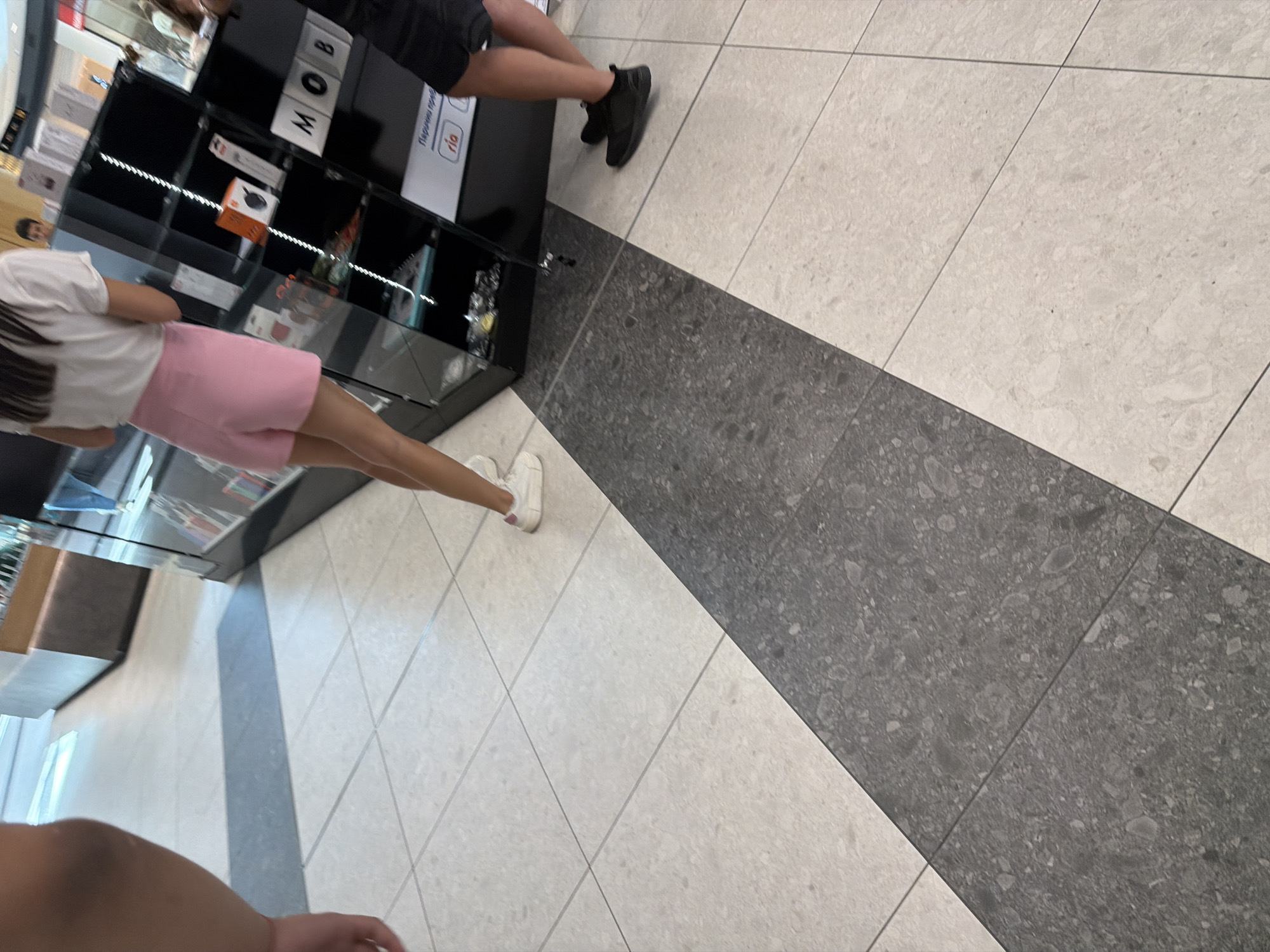 The image shows a section of a shopping mall with two individuals walking on a tiled floor. The floor is a mix of light and dark grey tiles, arranged in a diagonal pattern. In the background, there is a display of electronics and accessories within a black glass cabinet. One person is wearing pink shorts and a white t-shirt, while the other is wearing black shorts and a black t-shirt. The person in pink is facing away from the camera, and the person in black is partially visible from the side. The overall environment appears to be a retail space within a modern shopping complex. The image was taken in the city of Burgas, Bulgaria.