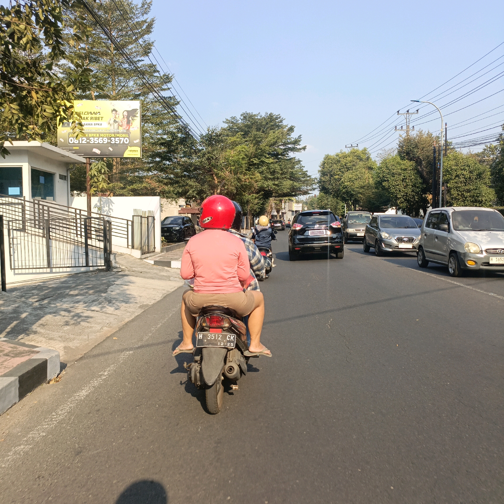 In Salatiga, Indonesia, a scene unfolds on a clear, sunny day along a paved road. The perspective is from behind a motorcycle, showing its rear and the passenger wearing a pink long-sleeved shirt and light brown shorts, with a red helmet on their head. The passenger's feet are bare, wearing flip-flops. Another person on a motorcycle, wearing a plaid shirt and a yellow helmet, is ahead. Several cars are on the road: a black SUV with license plate "H 1126 XH" in front, a silver car in the middle lane, and a silver car on the right. To the left of the road, a white building with a blue-tinted window is visible, bordered by a black metal fence and a ramp. A billboard with yellow and white text and images of people and money is mounted on poles near the building. Trees with green foliage line the sides of the road, and power lines crisscross overhead. The road has a white dashed line marking the lane and a curb with a concrete and red brick border on the left. Shadows from the trees and the motorcycles are cast on the road and sidewalk. The license plate of the motorcycle in the foreground is "H 3512 CK" with a date "12.25" below it.
