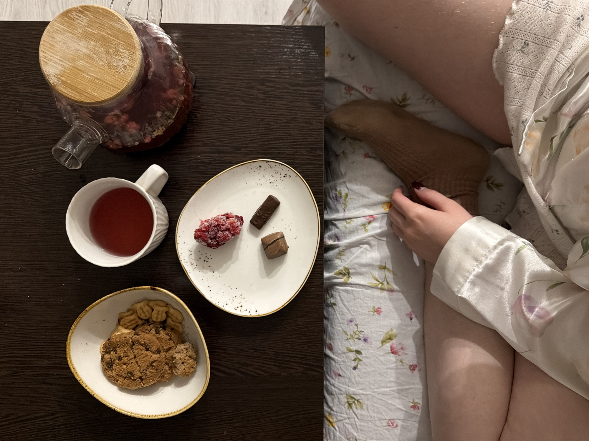 The image shows a still life of a meal on a dark wooden table next to a person lying in bed.
On the table, there is a glass teapot filled with a red-colored beverage and covered with a wooden lid. A white mug is positioned beside it, also filled with the same red drink. A white plate holds a cluster of red berries (possibly raspberries), and two pieces of dark chocolate. A small, off-white bowl contains cookies and animal crackers.
Beside the table is a person resting in bed. They are wearing a beige sock on one foot and covered with patterned white and pink floral bed linen. The person is partially covered by a white silky garment, possibly a pajama top or robe with floral patterns. A hand is visible near the sock.
There is no information to determine the location as Засечное, Russia.