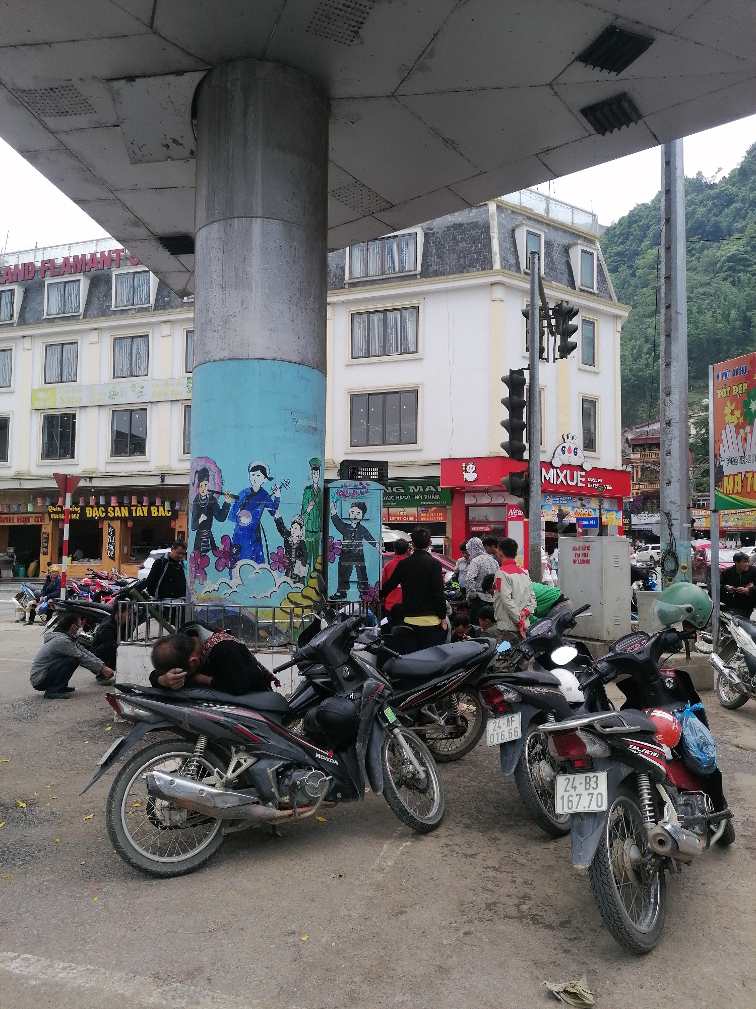 The image captures a scene in Sa Pa Ward, Vietnam, dominated by parked motorbikes beneath a concrete structure supported by a painted pillar. A person is resting their head on the seat of a motorbike. Other people are gathered in the area. Buildings with French architectural influences are in the background. Storefront signs such as "MIXUE" and "ĐẶC SẢN TÂY BẮC" are visible. A traffic light and a large advertisement poster are nearby.