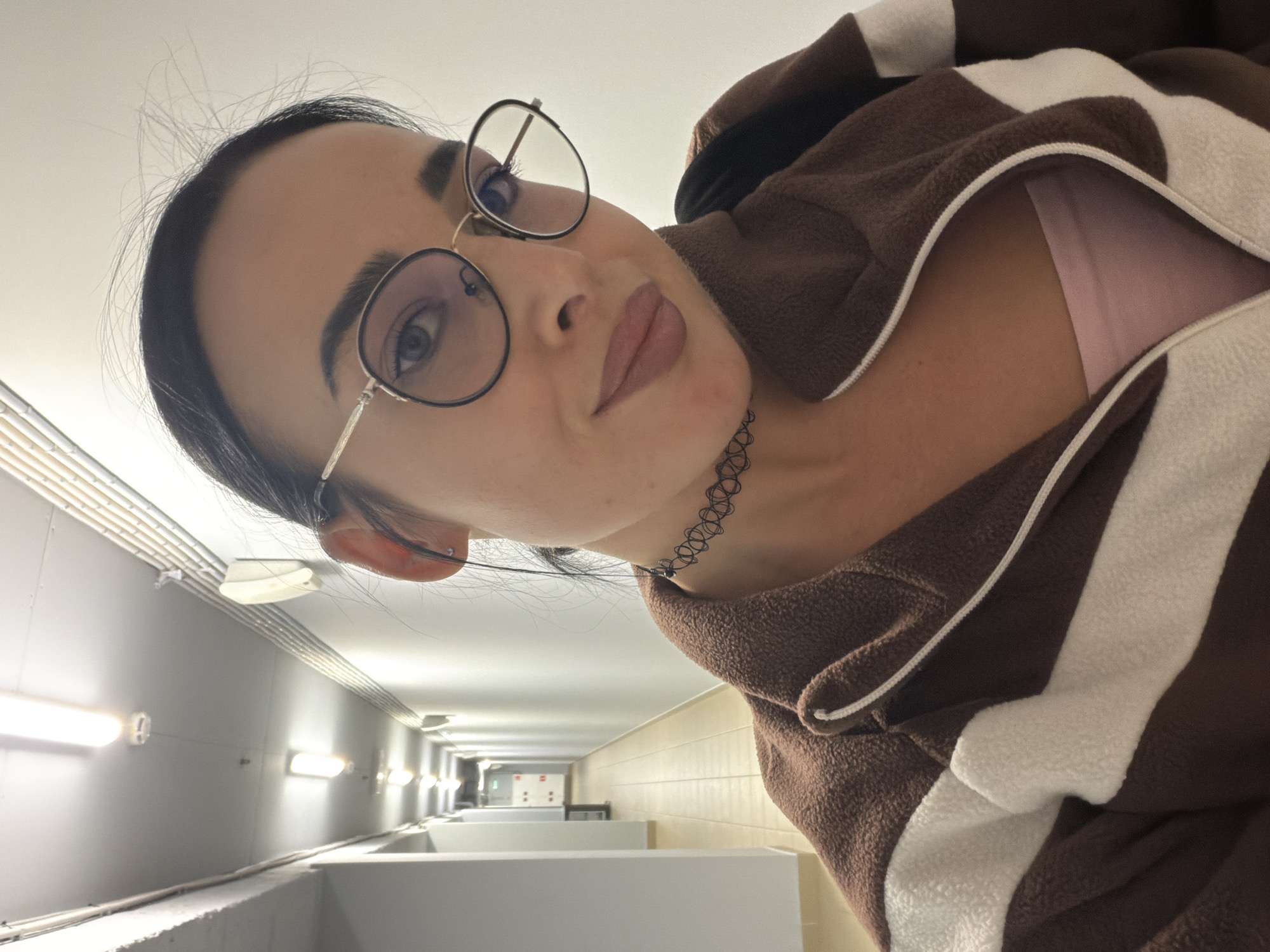 The image is a portrait of a young woman, likely a selfie, taken from a slightly low angle, showing her face and upper torso. She has dark hair, dark eyebrows, and is wearing round, thin-framed glasses. Her lips are full and appear to have a matte, neutral-toned lipstick. Around her neck is a black, patterned choker necklace. She is dressed in a brown and white striped hoodie, which appears to be made of a soft, fleece-like material. A glimpse of a light pink top is visible underneath the hoodie. Her expression is calm and neutral, with a direct gaze towards the camera.
The background depicts an indoor hallway or corridor with utilitarian design. The walls are light-colored, and fluorescent lights are mounted along them, creating a brightly lit space. Visible on the ceiling are some pipes or conduits. In the mid-ground of the background, there are several white, rectangular structures that resemble partitions or cubicles, suggesting a public or institutional setting like a changing room, public restroom, or a similar facility. The perspective of the hallway recedes into the distance, showing more of these structures and lights. No visible text is present in the image. The location is given as Saint Petersburg, Russia.