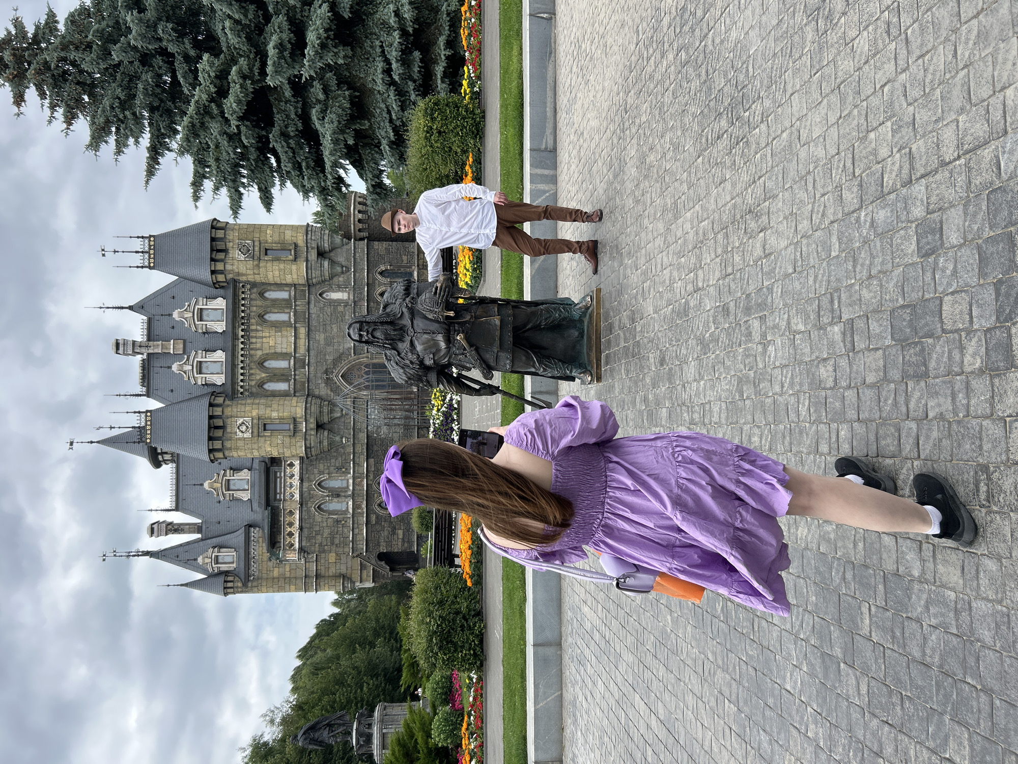 In Khryashchevka, Russia, a young woman in a purple dress is taking a photo of a young man posing next to a bronze statue of a medieval warrior. The man, wearing a white shirt and brown pants, stands with one hand resting on the statue's shoulder, holding a helmet. The statue depicts a bearded figure in armor with a sword. Behind them, a grand, castle-like building with multiple towers and grey spires stands under a cloudy sky. A large evergreen tree is visible to the right of the castle. The foreground is a paved courtyard with a pattern of grey stone blocks. Flower beds with colorful flowers are visible in the middle ground, leading up to the castle.