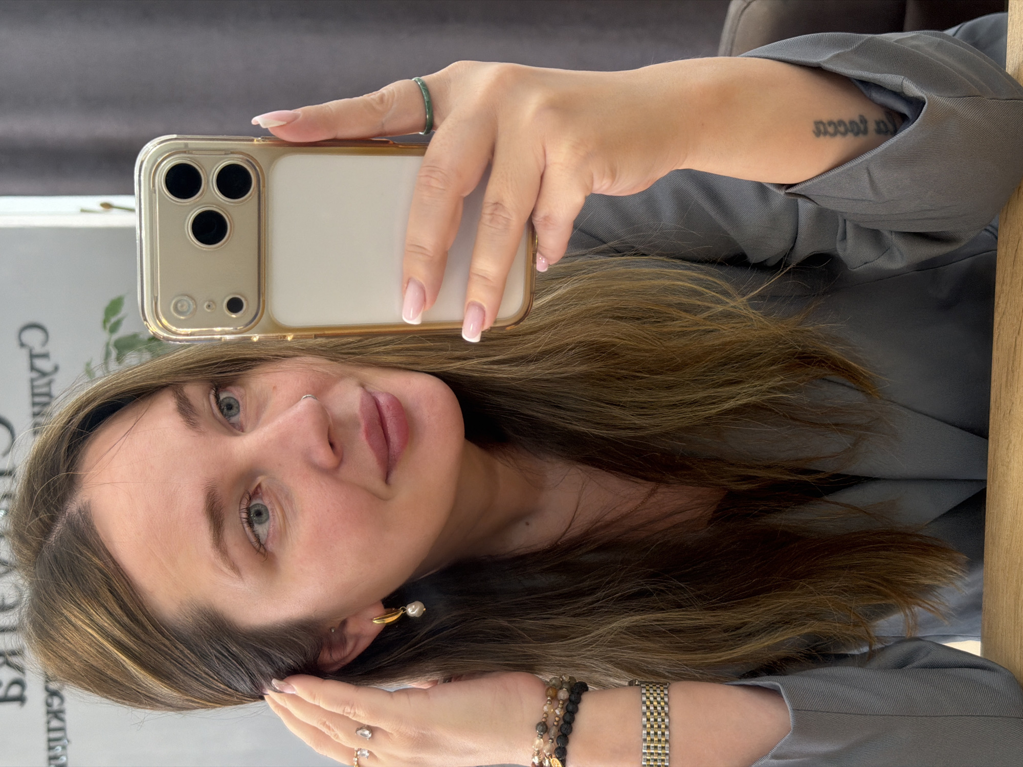 A woman with brown hair, light eyes, and a nose ring is depicted in a selfie, holding a gold-encased smartphone with her right hand. She is looking slightly to her right, with a subtle smile. Her left hand is raised, touching her hair near her ear, which displays a gold hoop earring with a pearl drop. On her left wrist, she wears a gold and silver two-tone watch and several beaded bracelets. A ring with a clear teardrop-shaped stone is on her left ring finger. Her right hand, holding the phone, shows a thin green band ring on her index finger. She is wearing a grey blazer, and a tattoo in dark script is visible on the inside of her right forearm. She is seated at a light wooden table. The background includes a light grey wall with reversed text, likely a reflection in a mirror, and a dark grey curtain to her right. The scene is located in the city of Chaikovsky, Russia, within what appears to be an indoor commercial setting such as a salon or office.