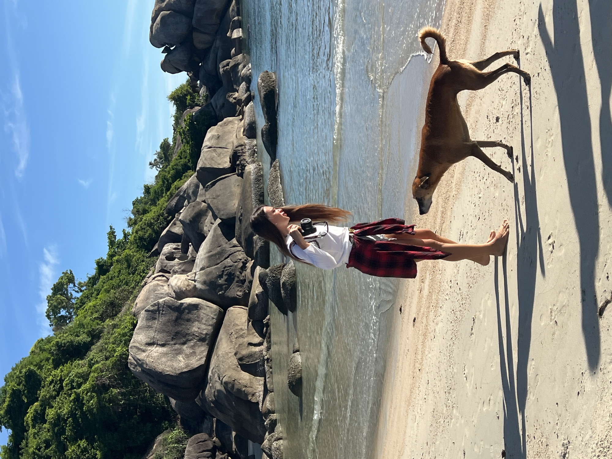In Baan Chaweng Noi, Thailand, a young woman with long dark hair stands barefoot on a sandy beach, facing a tan-colored dog. She is wearing a white long-sleeved shirt, denim shorts, and a red and black plaid shirt tied around her waist. She holds a camera in her right hand. The dog is facing the woman, with its tail curled upwards. The ocean is behind them, with clear, shallow water and visible rocks. In the background, large granite boulders and lush green vegetation cover a hillside, under a clear blue sky with a few wispy clouds. The sun casts long shadows on the sand from the woman and the dog.