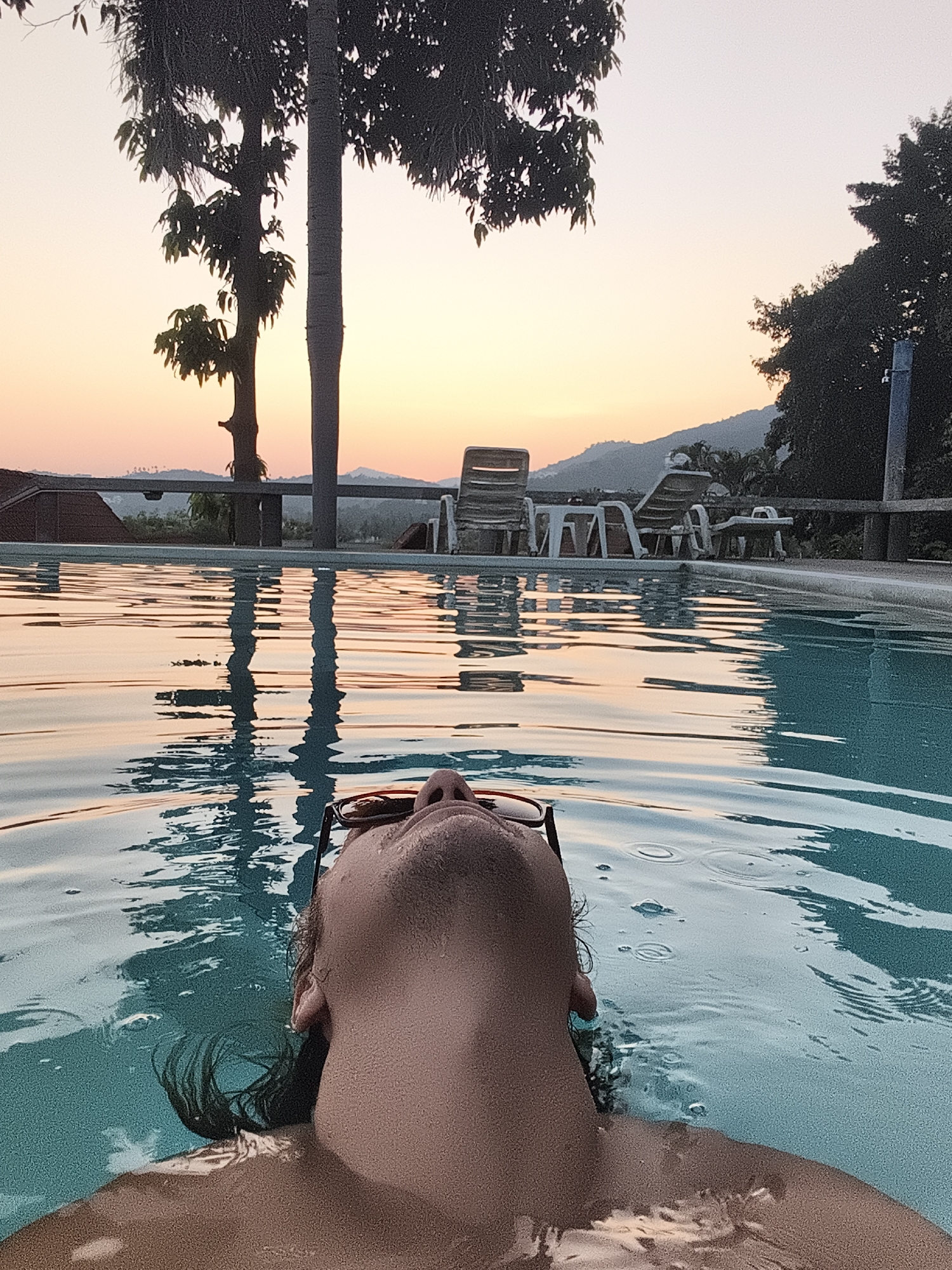 A person lies on their back, floating in a swimming pool. The person is wearing sunglasses and the pool water comes up to their neck. Behind the person, at the edge of the pool, are lawn chairs and a tree. Beyond that, the sky is a gradient of oranges and yellows, indicating a sunset. The location is บ้านบางรักษ์, Thailand.