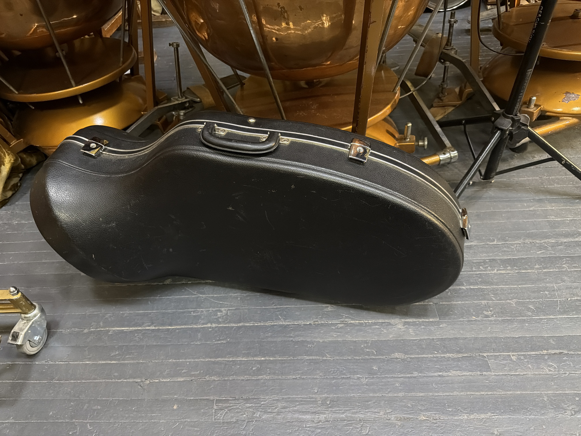 An overhead shot captures a black, hard-shell instrument case resting on a dark gray floor, surrounded by large copper timpani drums and various stands. The instrument case, shaped to house a brass instrument like a French horn or a small tuba/euphonium, is positioned centrally, angled towards the left. It features a sturdy handle, silver-colored latches, and a thin white or silver stripe running along its top edge, showing some scuffs and wear.
In the background and partially obscured, several timpani drums are visible. These large, round, copper-colored instruments are supported by metal stands, some with gold-painted bases. The drum bowls exhibit reflections and some minor imperfections, consistent with use. A black, multi-legged stand, possibly for percussion or a microphone, is positioned to the right of the case. Partially visible text on this stand reads "PERCUSSION".
The floor is a dark, possibly charcoal gray, with horizontal grooves or lines suggesting planks or a patterned surface, appearing worn in places. In the bottom left corner, a single metal wheel attached to a golden or brass-colored frame is visible, indicating other equipment is nearby. The overall setting appears to be an indoor storage area, rehearsal room, or backstage space for a musical ensemble, with no human activity taking place. The lighting suggests an indoor environment.