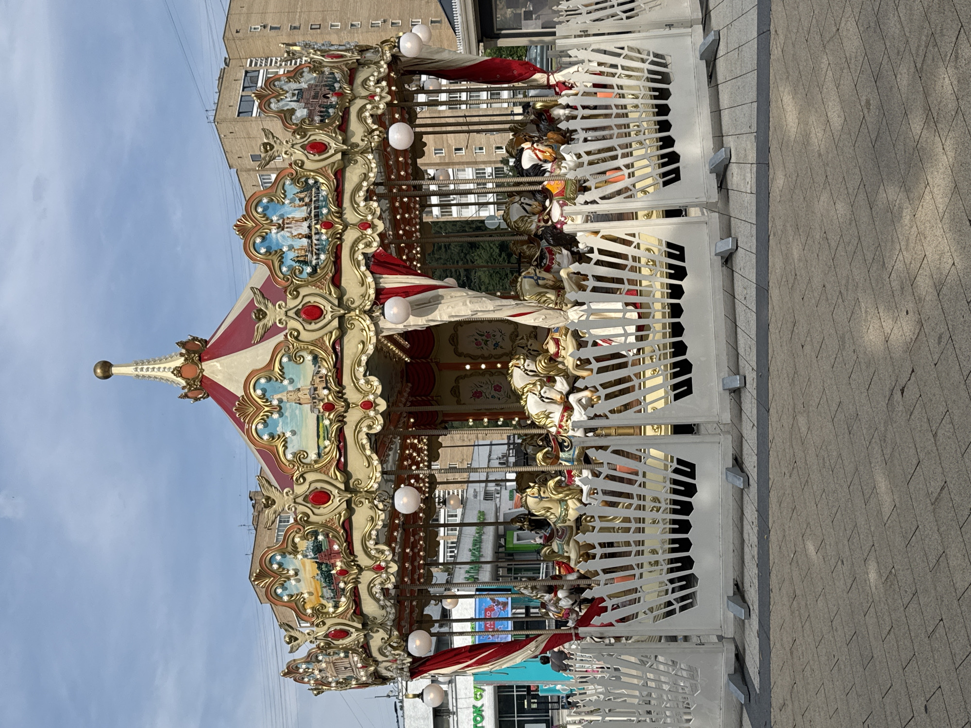 A carousel is set up in an outdoor urban environment with buildings visible in the background. The carousel is elaborately decorated with red and gold accents, ornate carvings, and painted murals depicting cityscapes. White horses, adorned with colorful saddles, are positioned around the carousel. The carousel is surrounded by a white picket fence with decorative cutouts. The ground is paved with gray bricks. The sky is partly cloudy. A few people are visible near the carousel, including one person in the foreground to the left looking at the carousel, and another person partially visible in the background to the right. The setting appears to be a public space in Zelenograd, Russia, given the context provided.