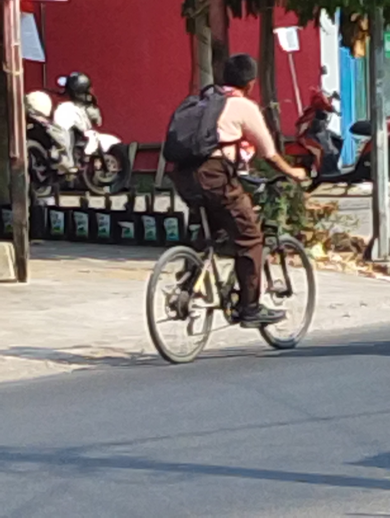 The image shows a person riding a bicycle on a street in Manaruwi, Indonesia. The person is wearing a backpack and brown pants. They are facing away from the camera and appear to be pedaling the bicycle. In the background, there are several motorcycles parked. The environment appears to be an urban setting with buildings and trees. The lighting suggests it is daytime.