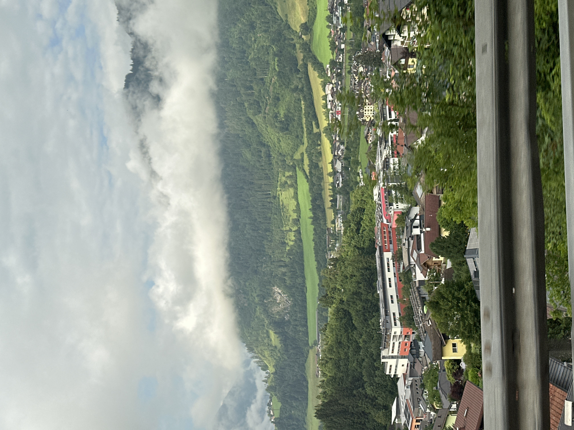 A hazy, overcast sky with patches of blue hovers over a lush green mountain range dotted with evergreen trees. The lower slopes are covered in dense forest, transitioning into rolling green fields. A town is nestled in the valley below, characterized by a mix of residential homes with red and brown roofs and larger, modern buildings with white, red, and grey facades. A metal guardrail is visible in the foreground. The scene depicts a rural town in Rettenstein, Austria, under a cloudy mountain sky.